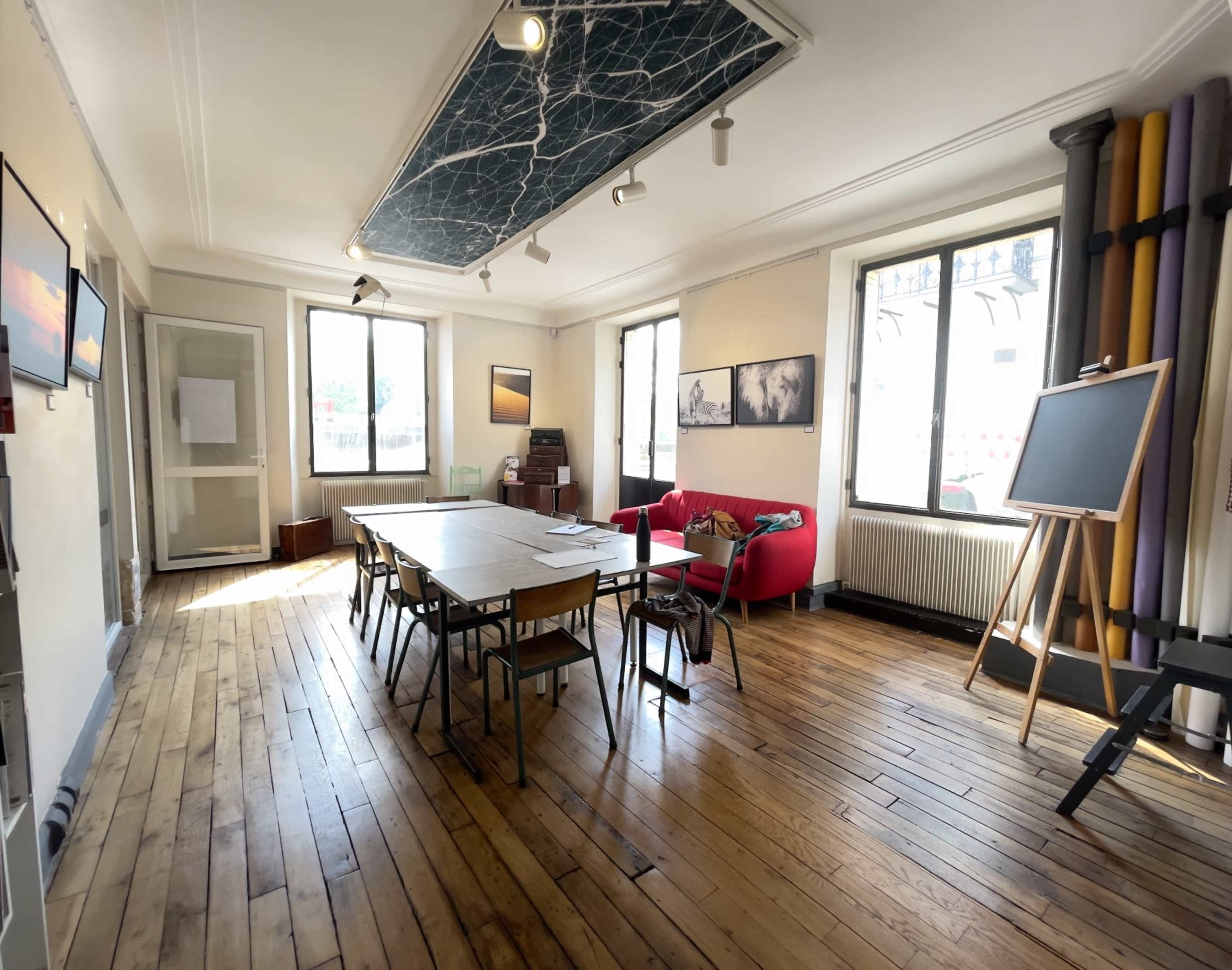 A bright meeting room with wooden floors, a long table surrounded by chairs, a red couch, large windows, and a chalkboard.