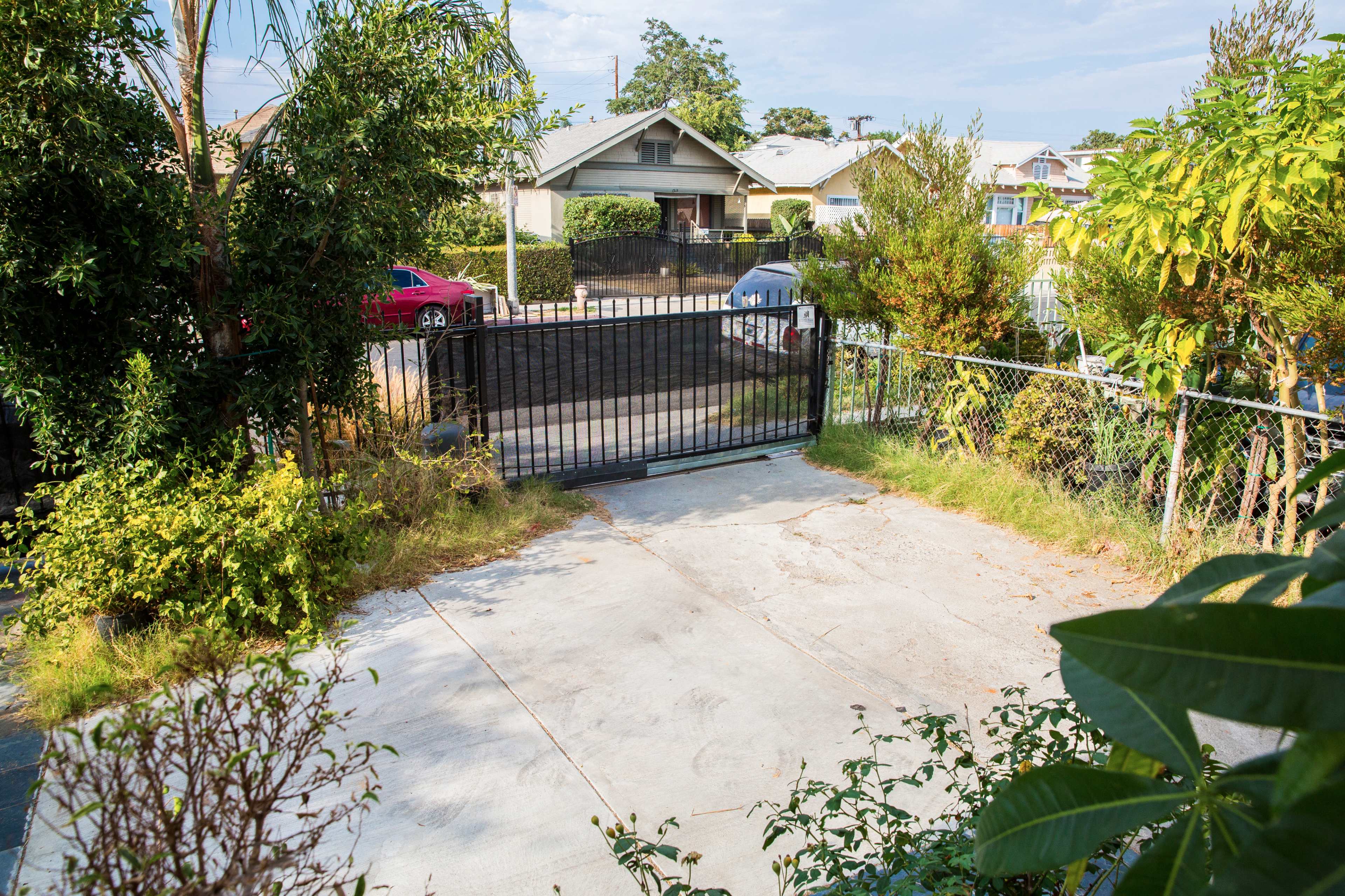The image shows an empty driveway with a gated entrance, surrounded by greenery and facing a residential street.