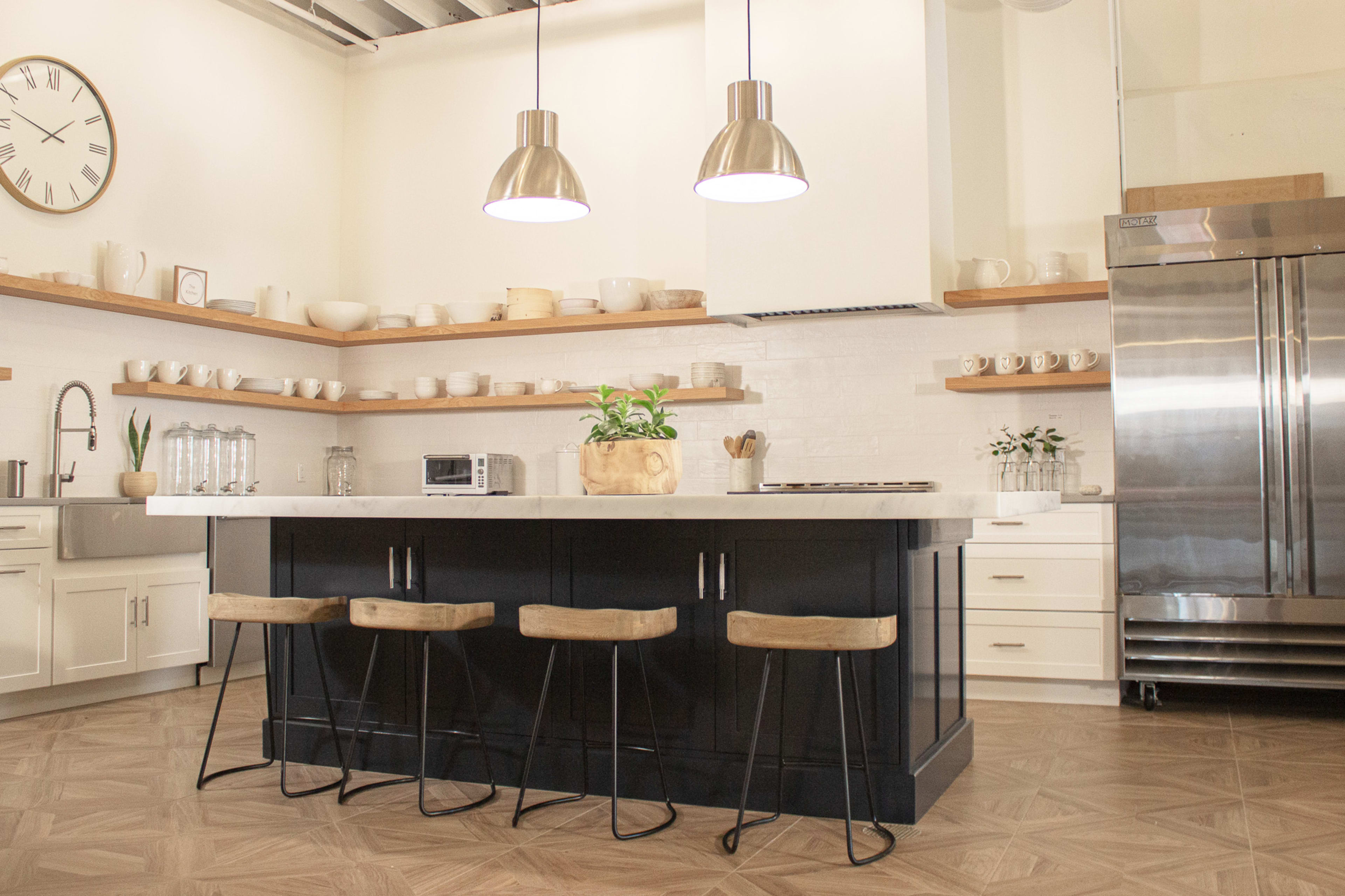 The image shows a modern kitchen featuring a large island with three wooden stools, stainless steel appliances, and shelves stocked with dishware.