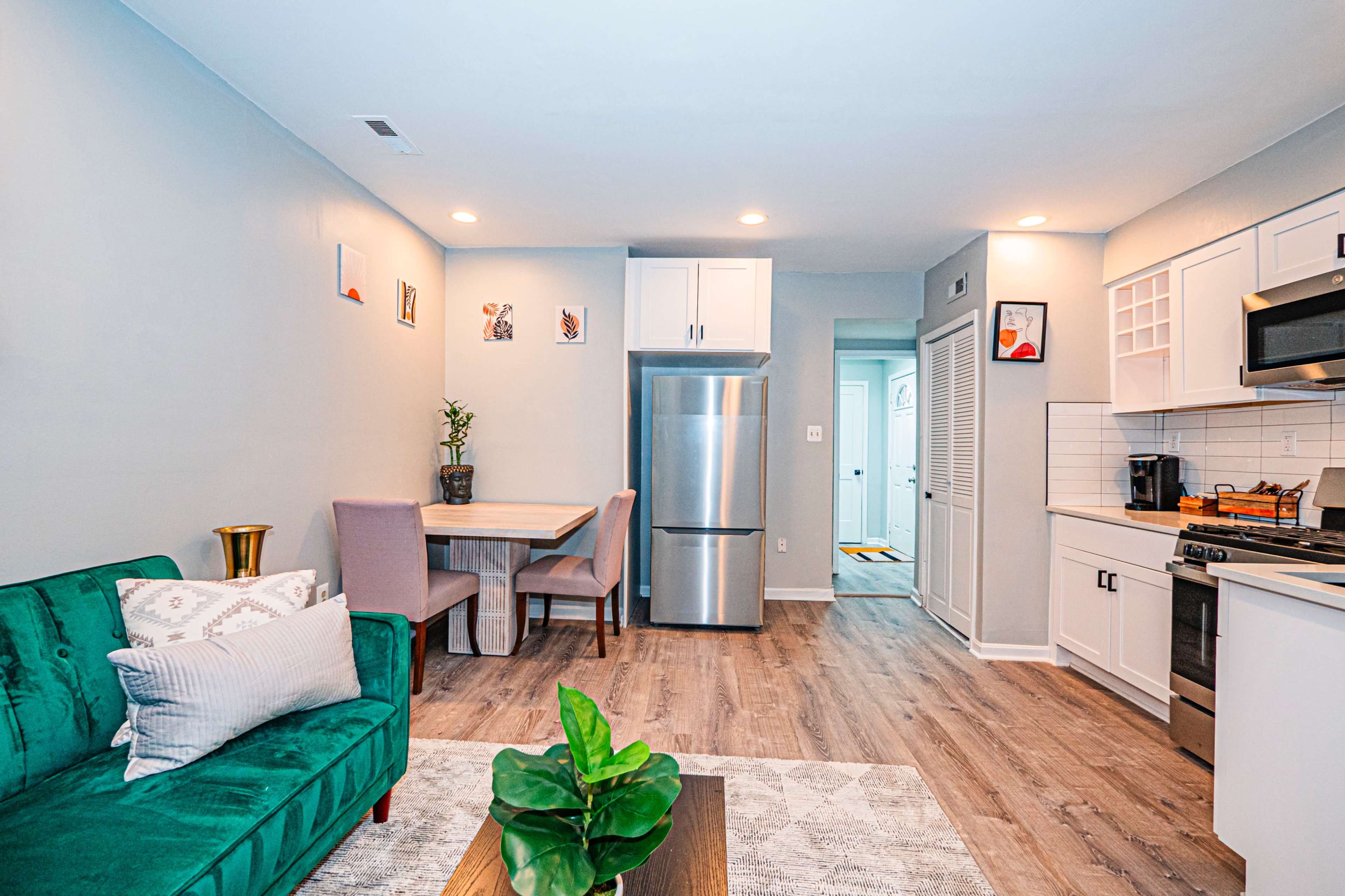 The image shows a modern kitchen and living area with a green sofa, a dining table with two chairs, stainless steel appliances, and light-colored cabinetry.