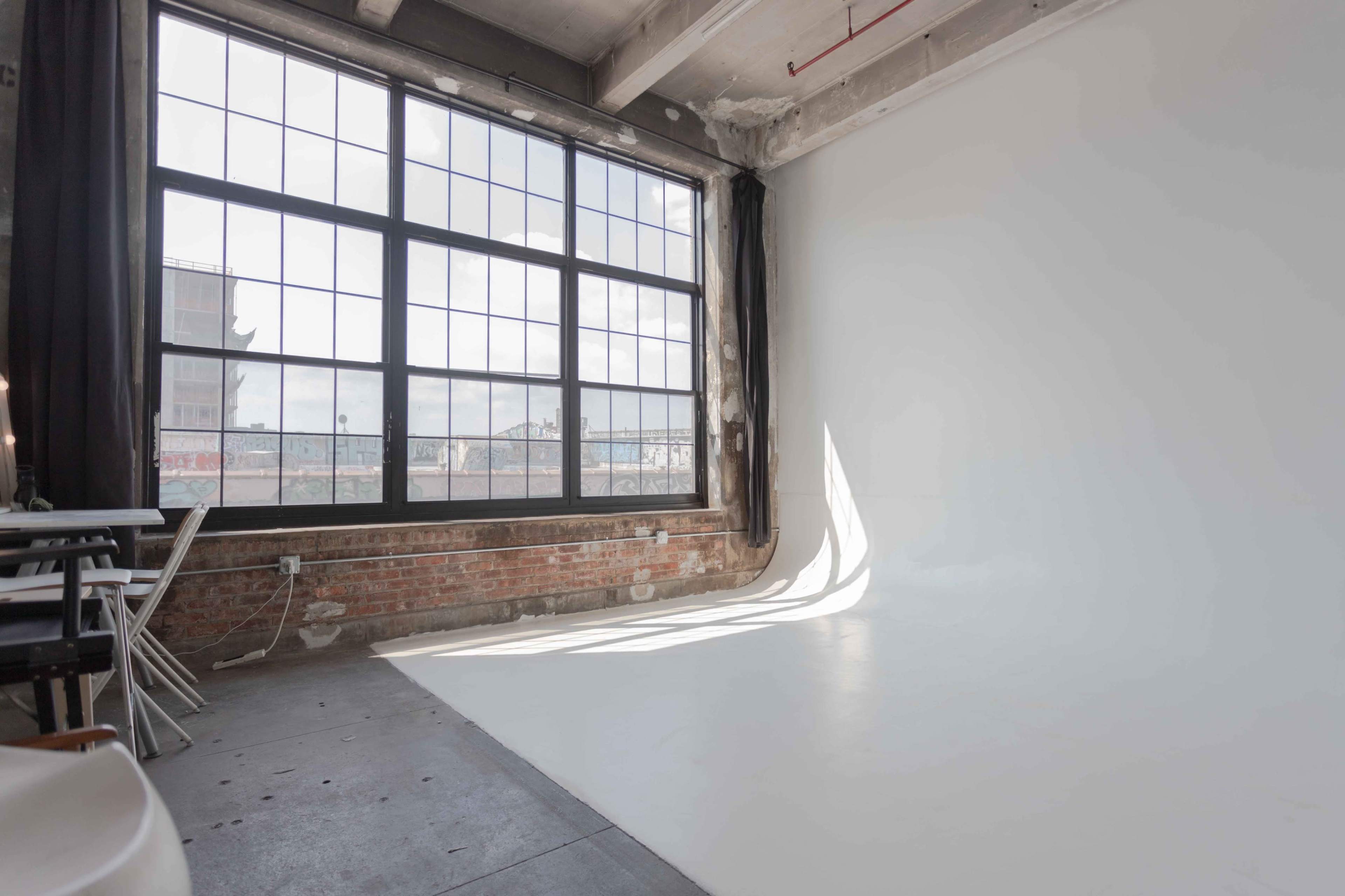 A spacious room with large industrial windows, featuring a white backdrop and exposed brick walls.