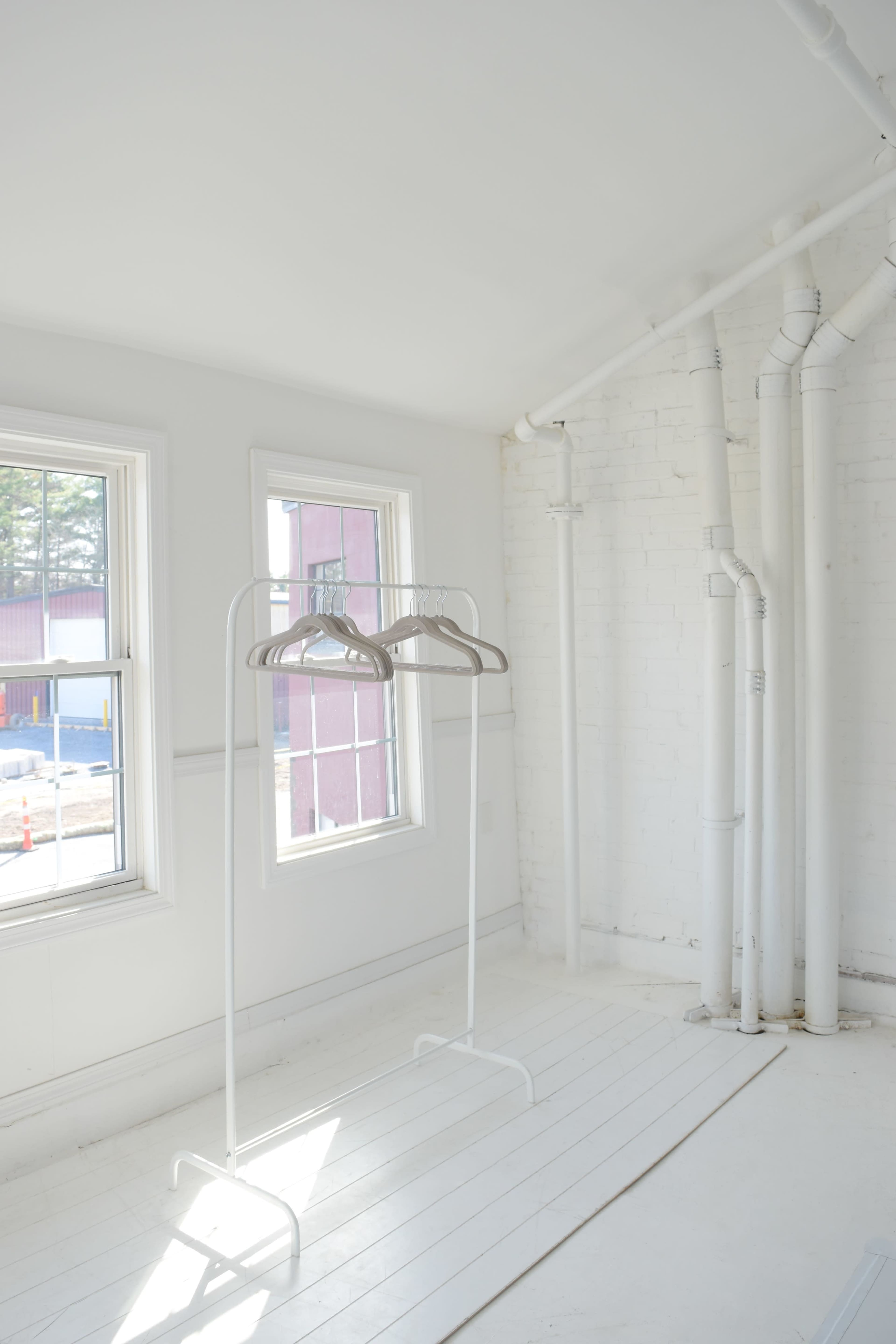 The image shows a bright, empty room with two white clothing racks and exposed pipes along the walls.