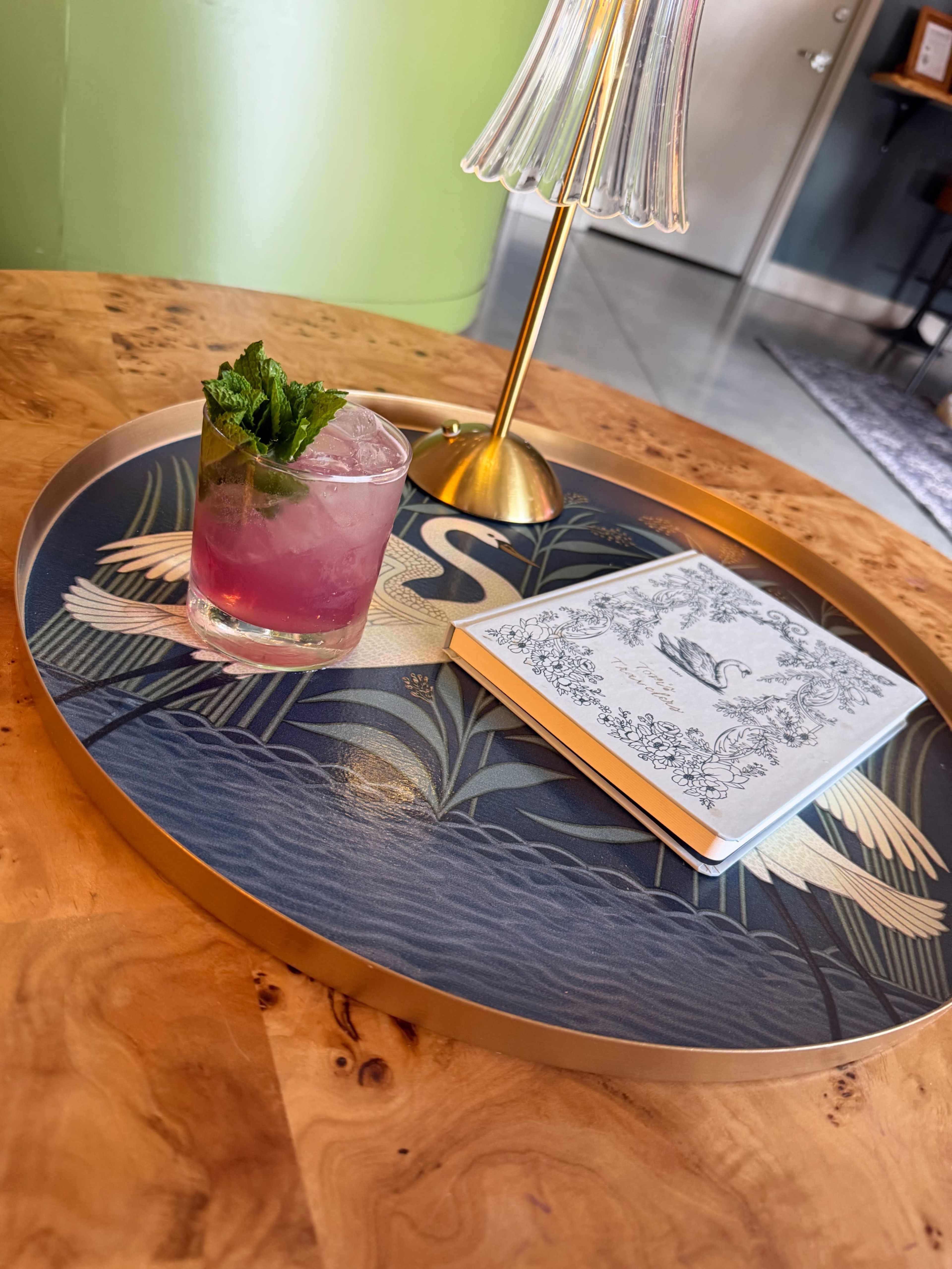 A cocktail with mint leaves sits on a decorative tray alongside a closed notebook in a well-lit indoor setting.