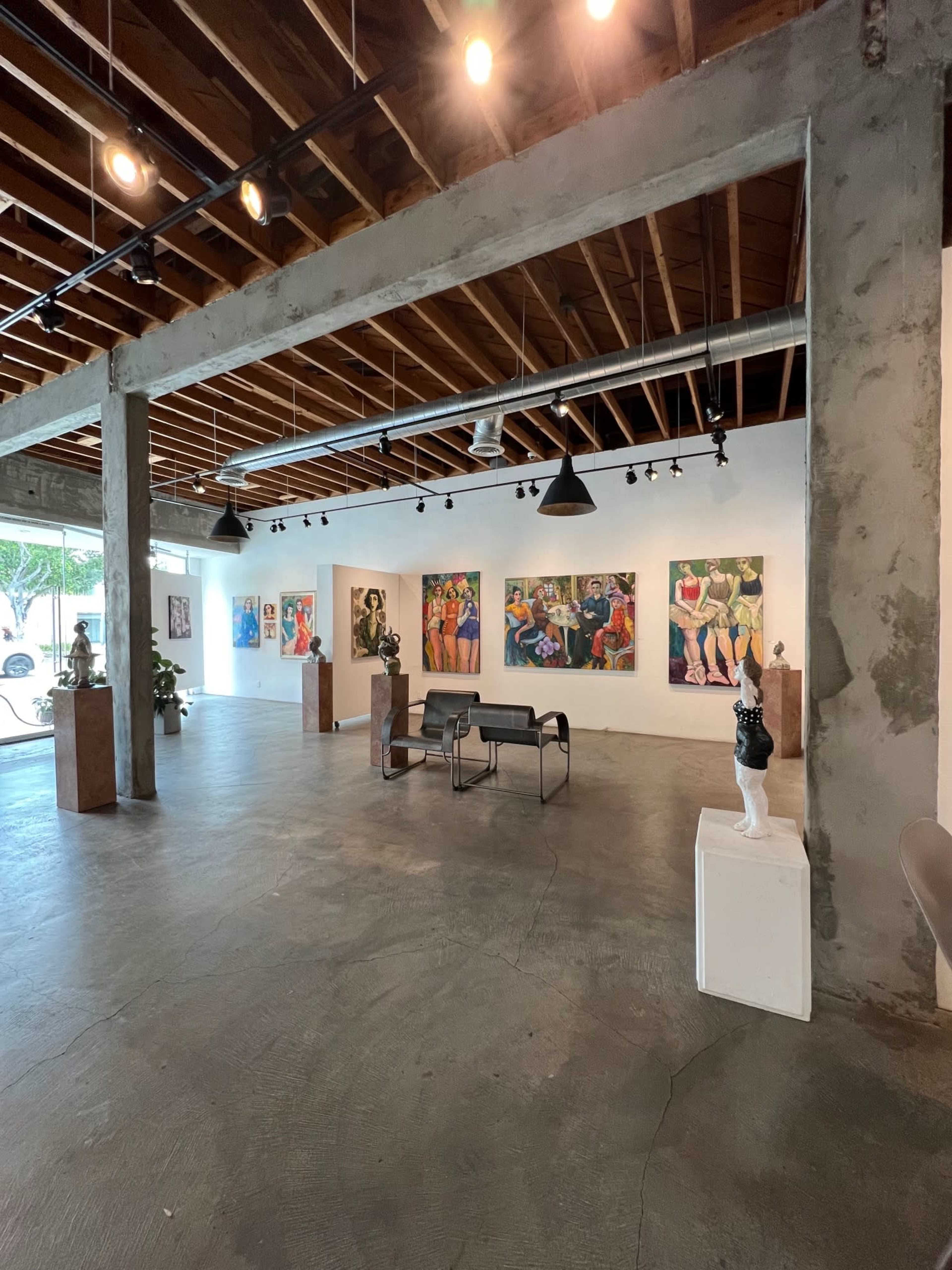 An art gallery with various paintings on the walls and sculptures displayed on pedestals, featuring a concrete floor and exposed wooden beams.