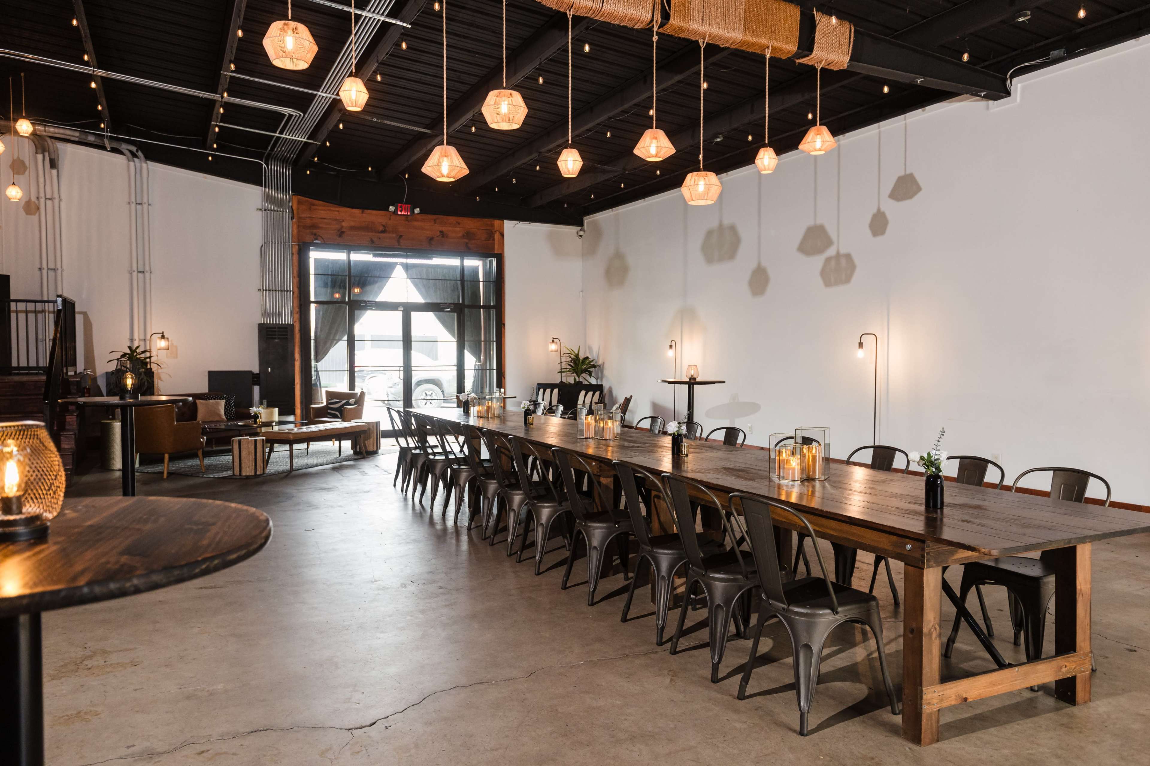The image shows a spacious dining area with a long wooden table, metal chairs, and warm hanging lights illuminating the neutral-toned space.