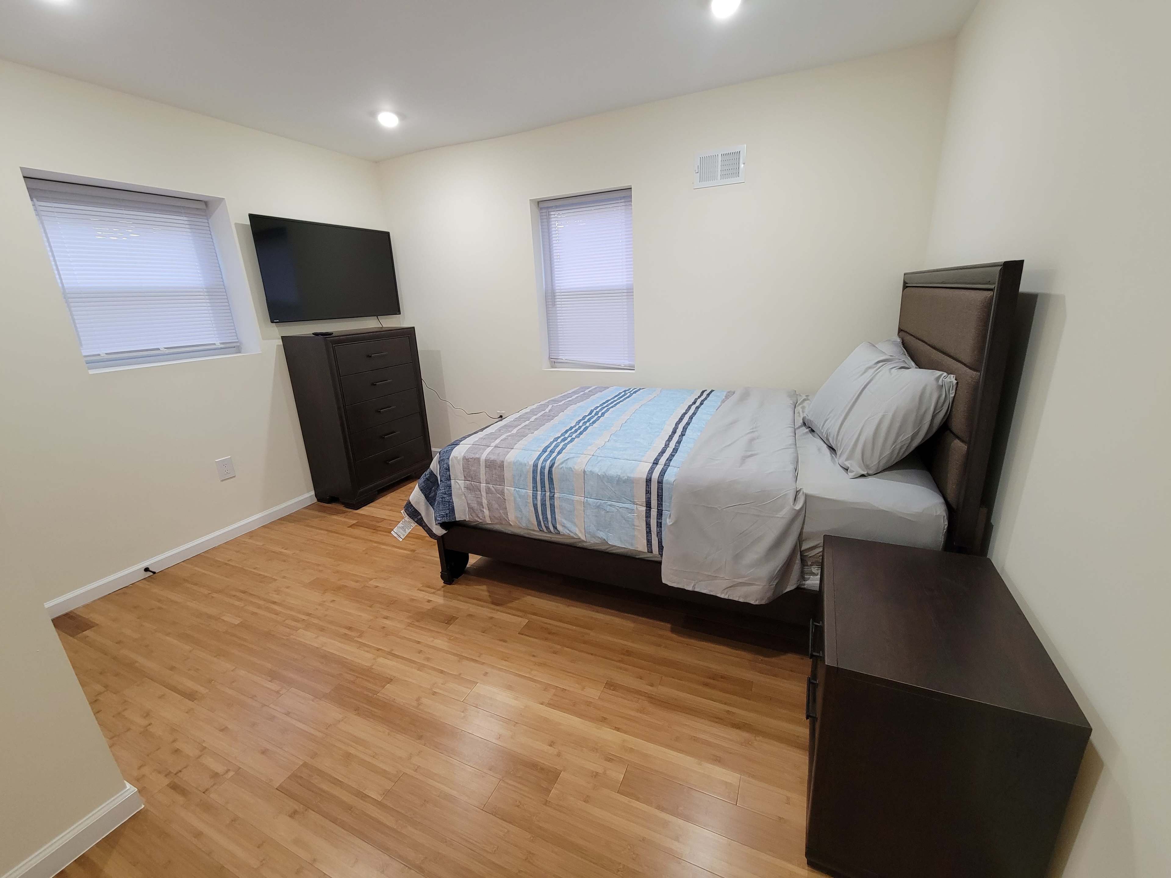 A simple bedroom features a bed with a striped blanket, a dresser, a nightstand, and a television mounted on the wall.