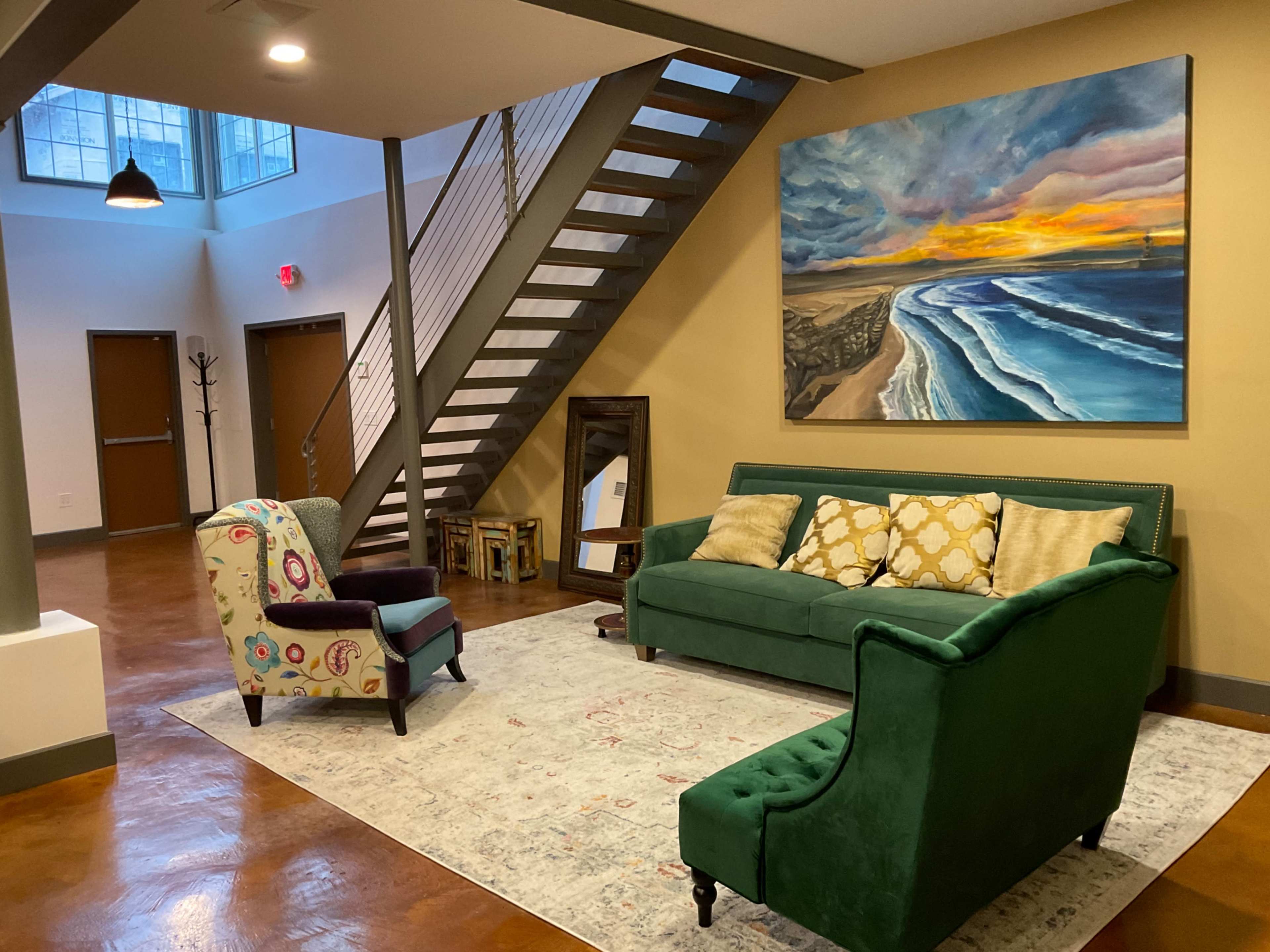The image shows a modern living area with a green couch, a patterned chair, and a large painting of a seascape on the wall, along with a staircase in the background.