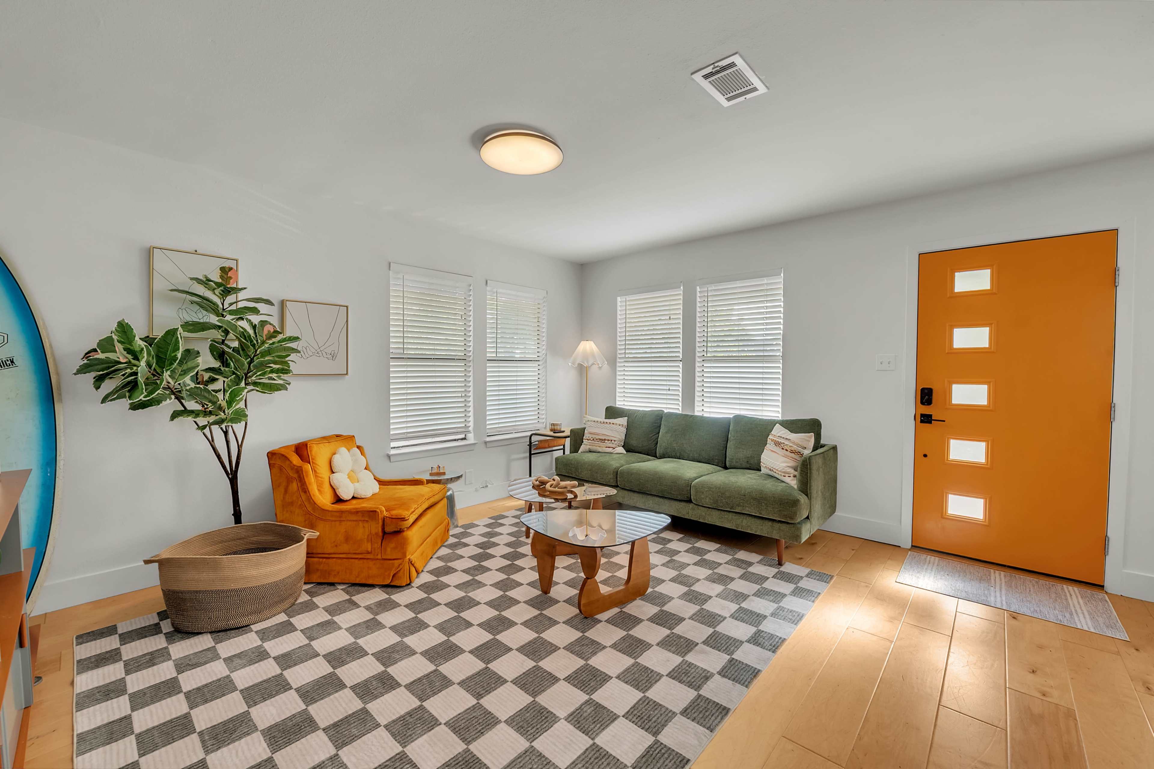 The image shows a living room featuring a green sofa, a yellow armchair, a checkered rug, a round coffee table, and an orange front door with square windows.
