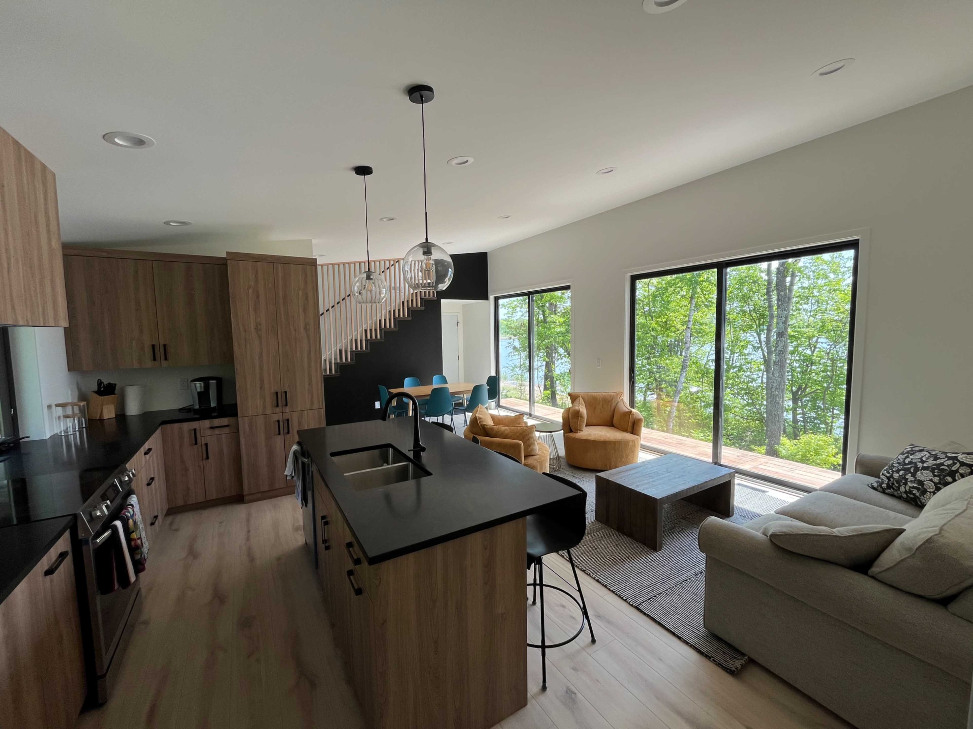 A modern open-concept kitchen and living area features wooden cabinetry, stainless steel appliances, and large windows overlooking a scenic view of trees and water.