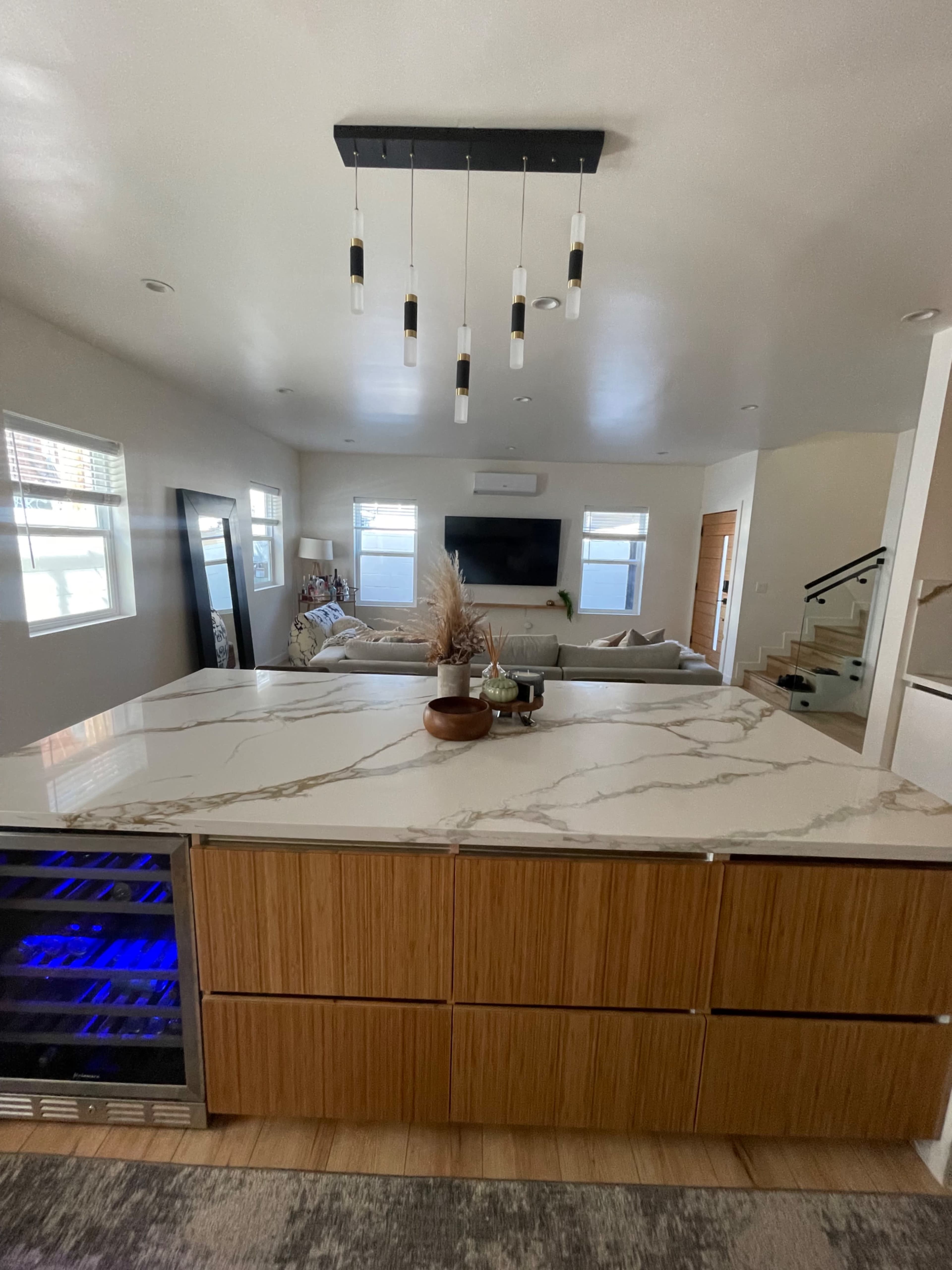 A modern kitchen features a marble countertop island with under-counter lighting, facing a living area with a sectional sofa and a mounted television.