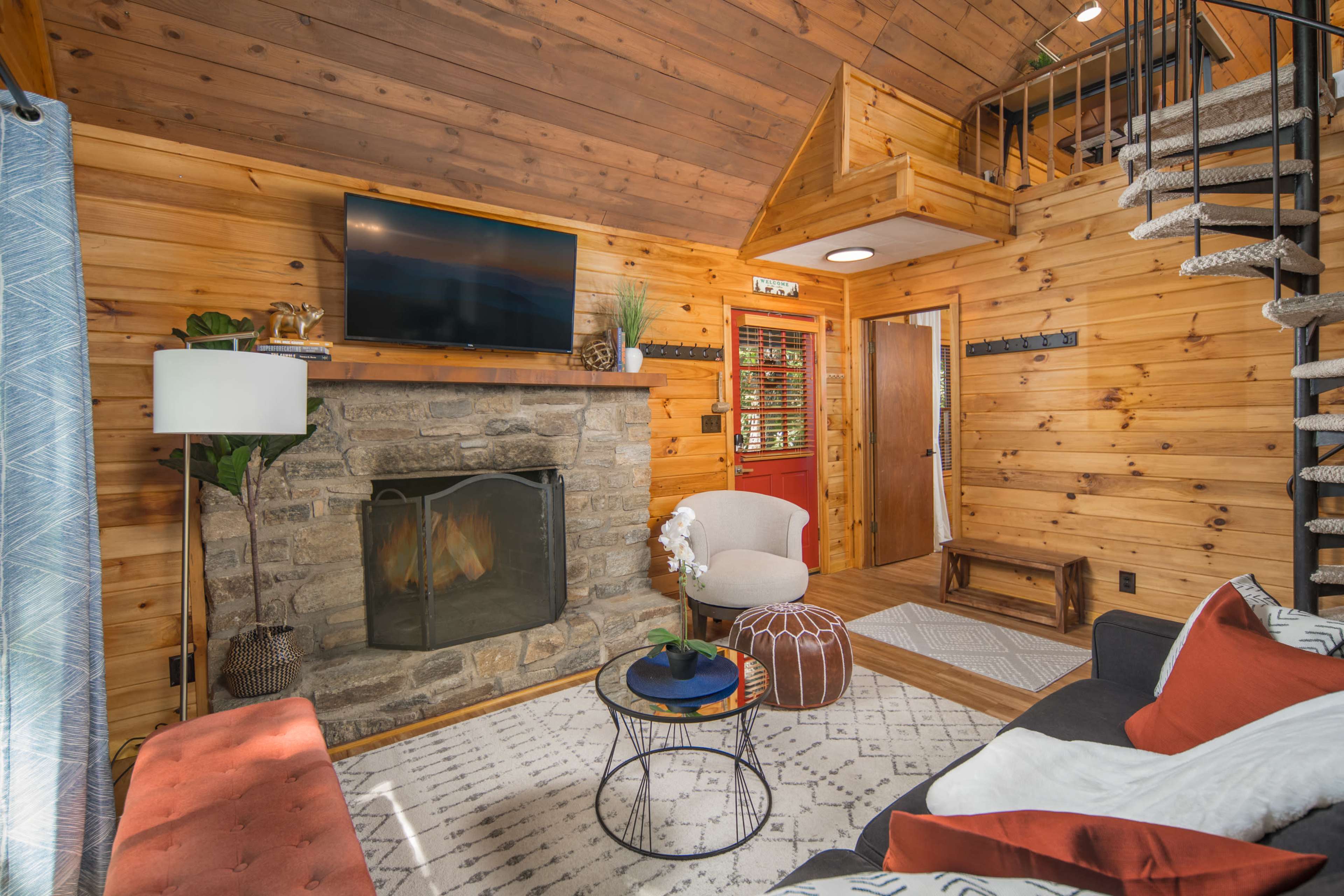 A cozy, wood-paneled living room with a stone fireplace, a television mounted above it, and a spiral staircase leading to an upper level.