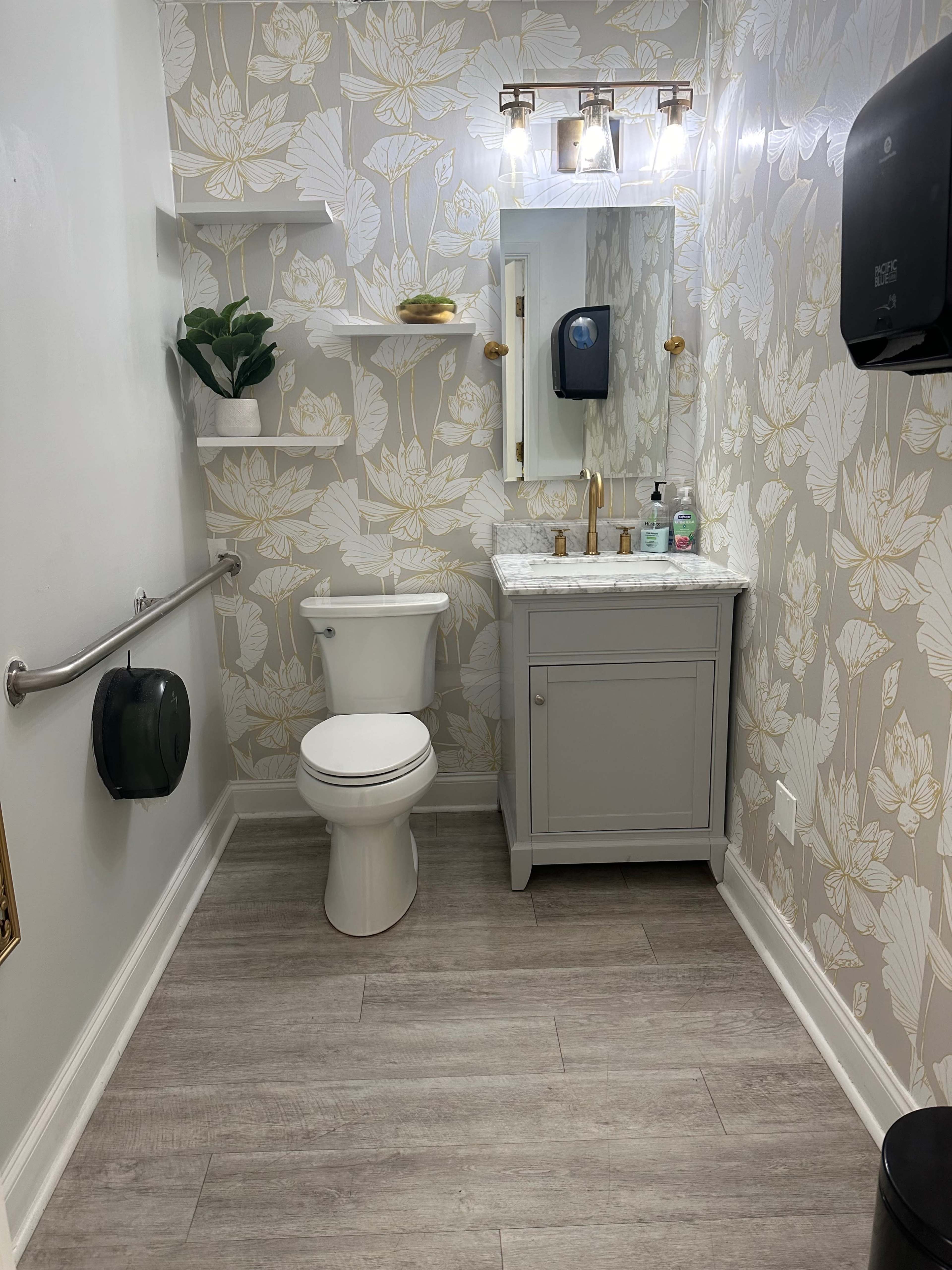 The image shows a clean bathroom featuring a white toilet, a sink with a marble countertop, wall shelves, and floral wallpaper.