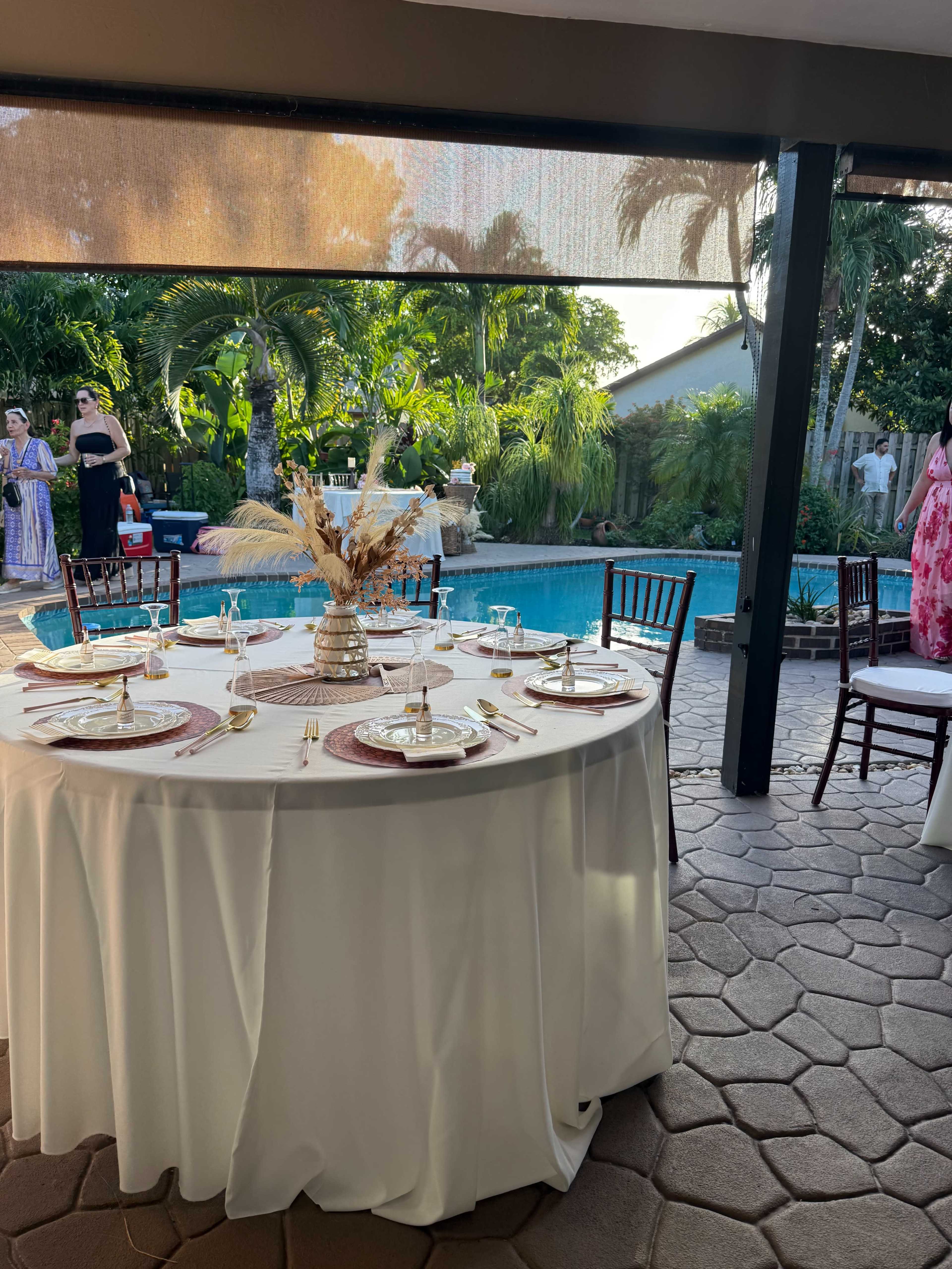 A round dining table is set with elegant tableware near a swimming pool, surrounded by lush greenery.
