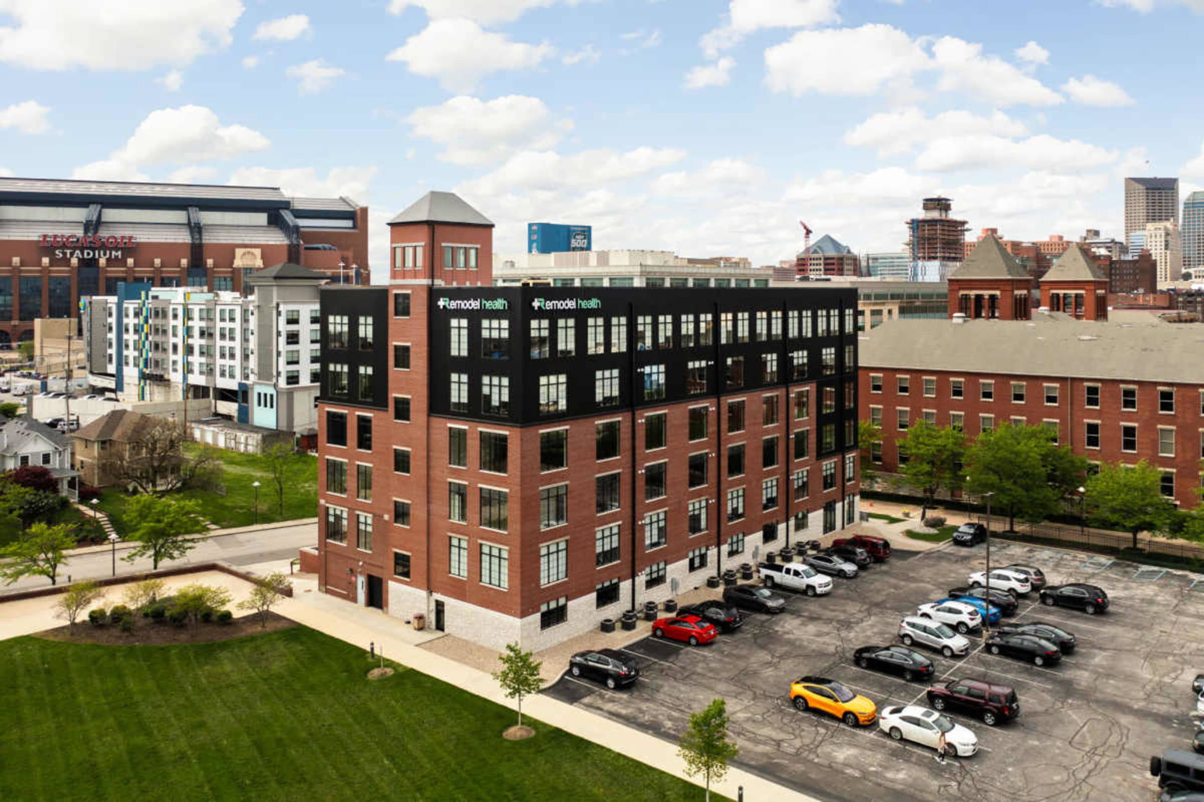 The image shows a multi-story brick building with a modern facade, surrounded by green grass and a parking lot, with a stadium and other buildings in the background.