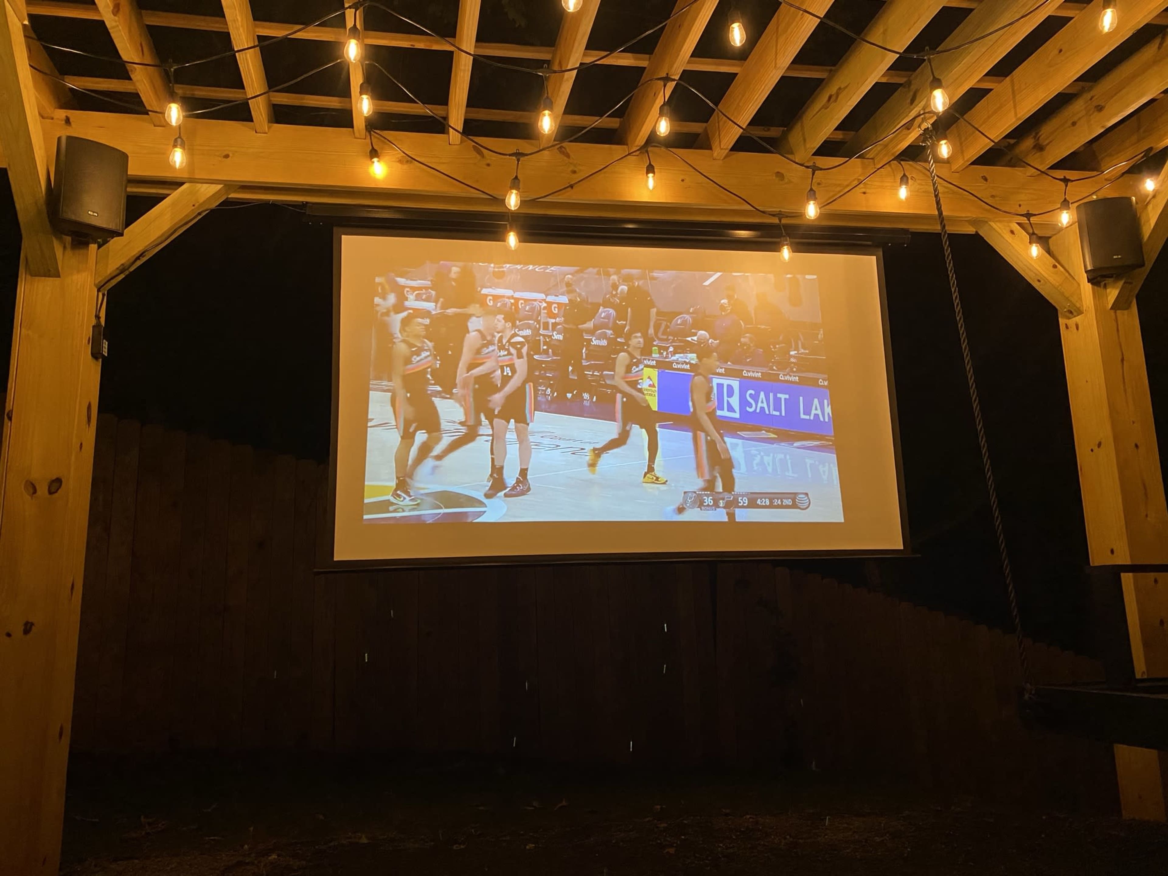 A basketball game is being displayed on a screen in an outdoor pavilion illuminated by string lights.