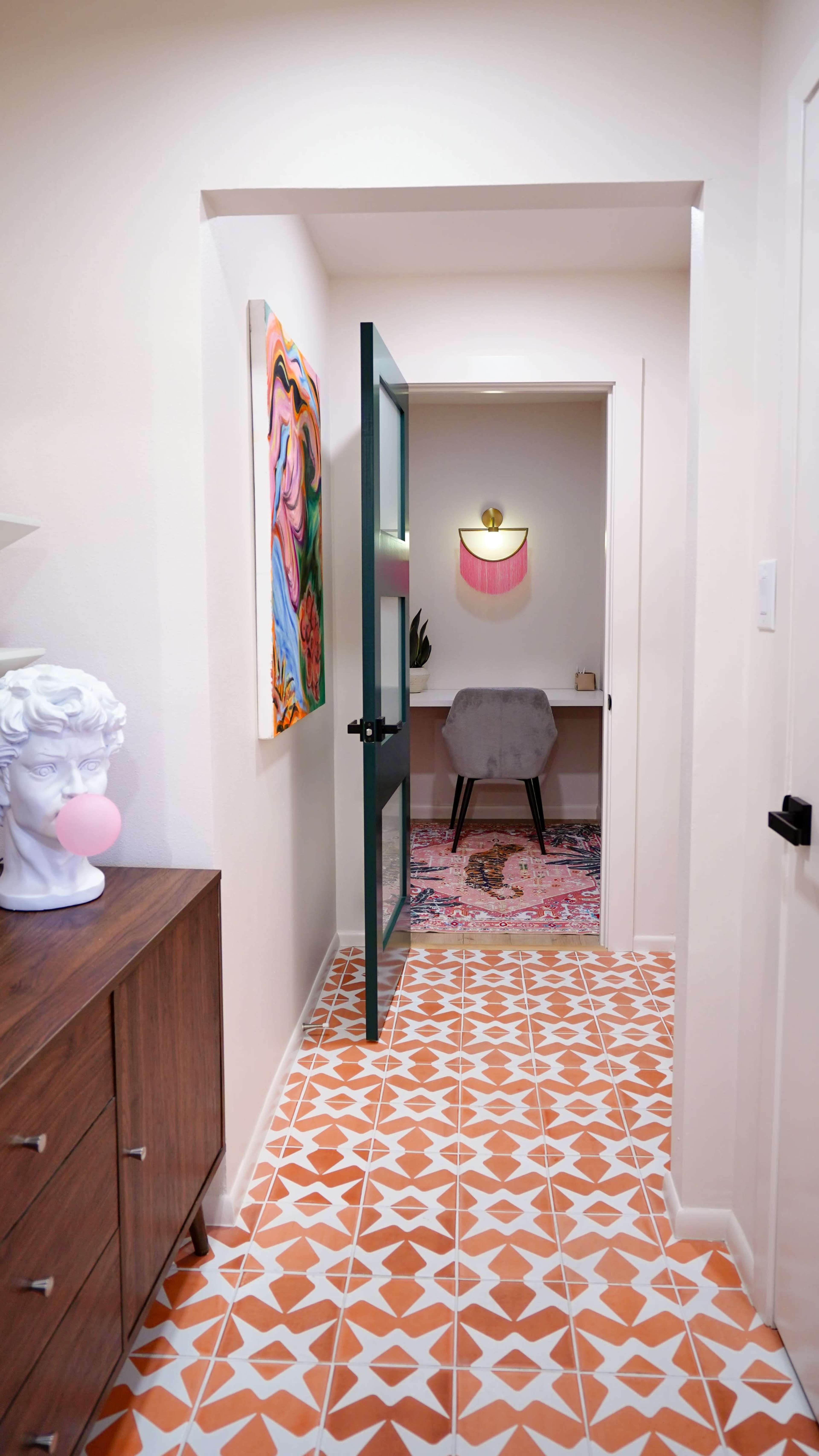A bright hallway features a green door leading to a small workspace adorned with colorful artwork and a patterned rug.