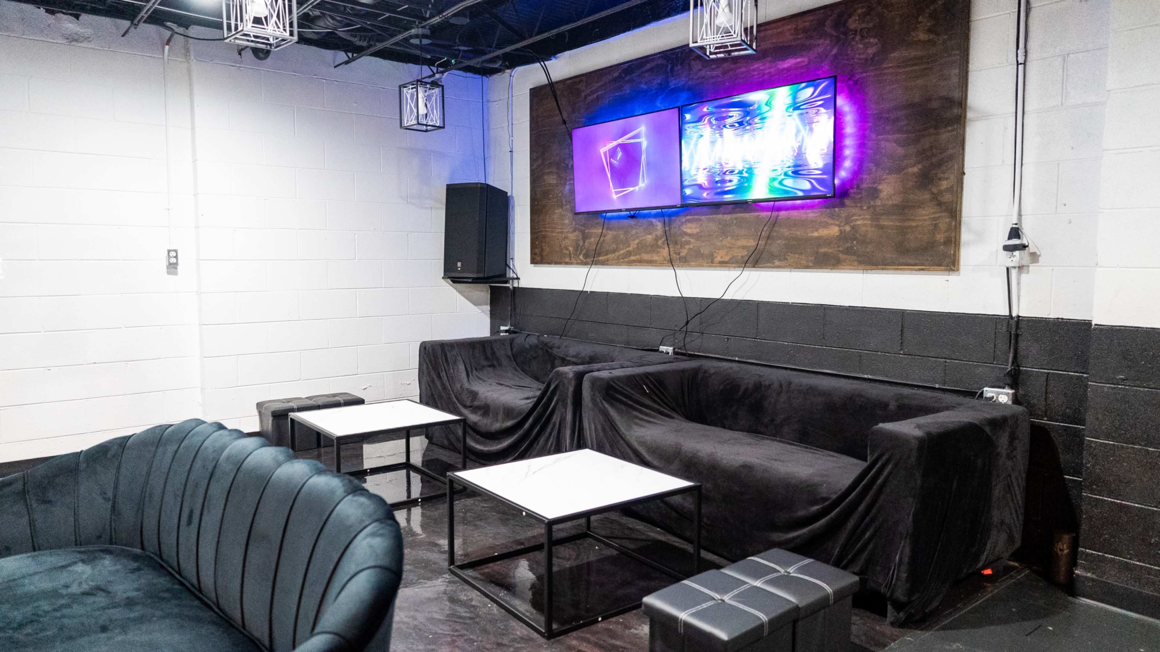 The image shows a lounge area with black sofas, white coffee tables, and a wall-mounted screen displaying colorful graphics.