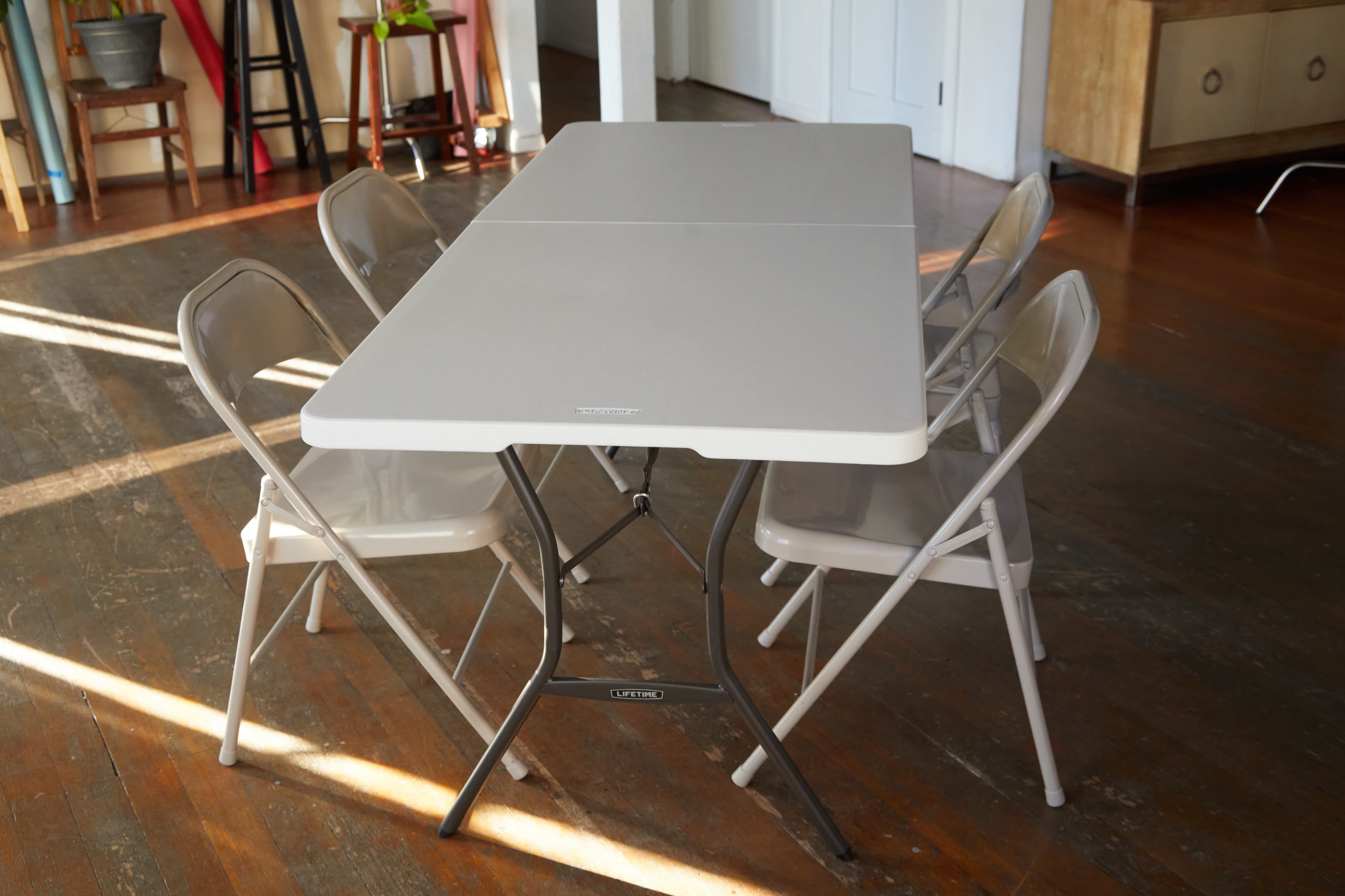 A rectangular folding table is surrounded by four folding chairs in a well-lit room with wooden flooring.