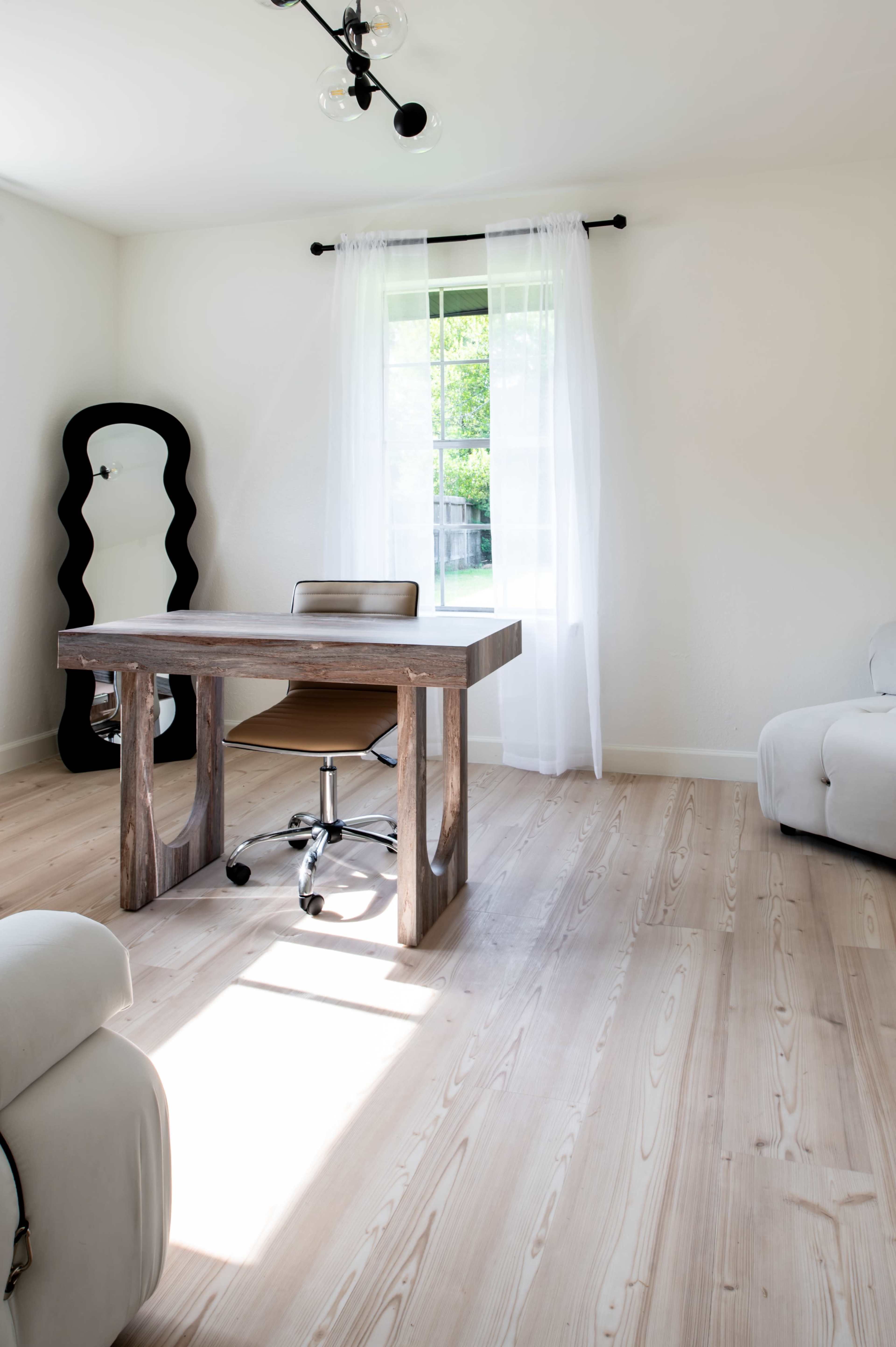 The image shows a minimalist office space featuring a wooden desk, an ergonomic chair, and a large mirror beside a window with sheer curtains, all on light-colored wooden flooring.