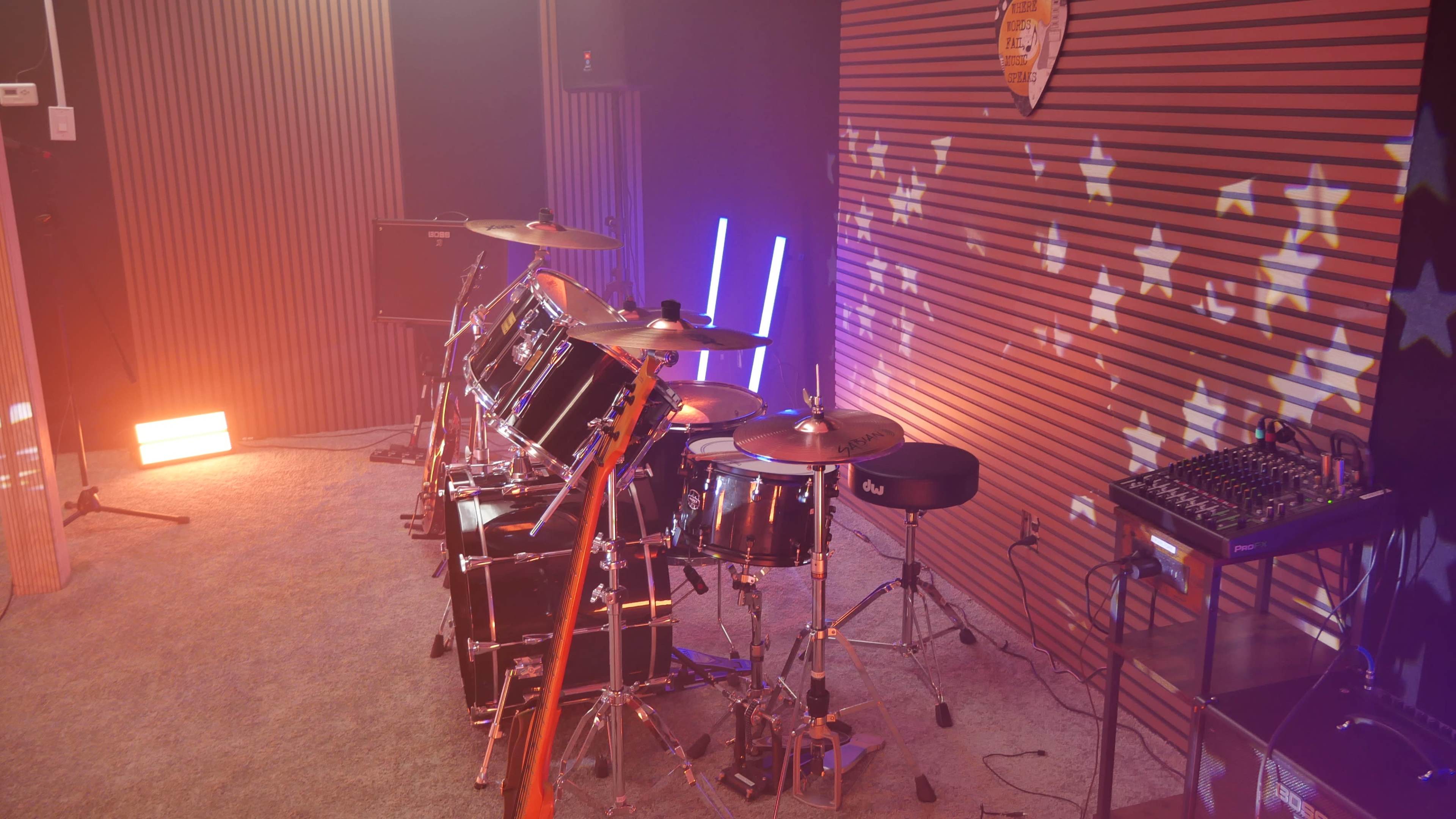 A drum set is positioned in a music studio with colorful lighting and sound equipment in the background.