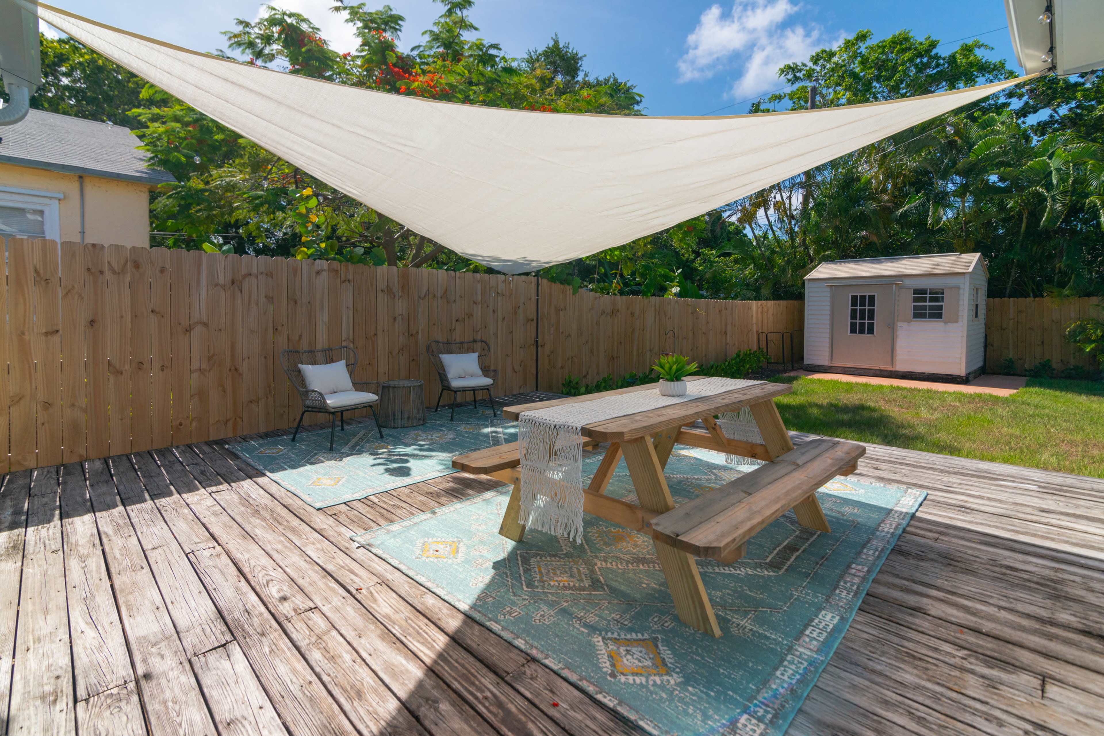 A shaded outdoor patio features a wooden table and chairs on a rug, surrounded by a wooden fence and greenery.