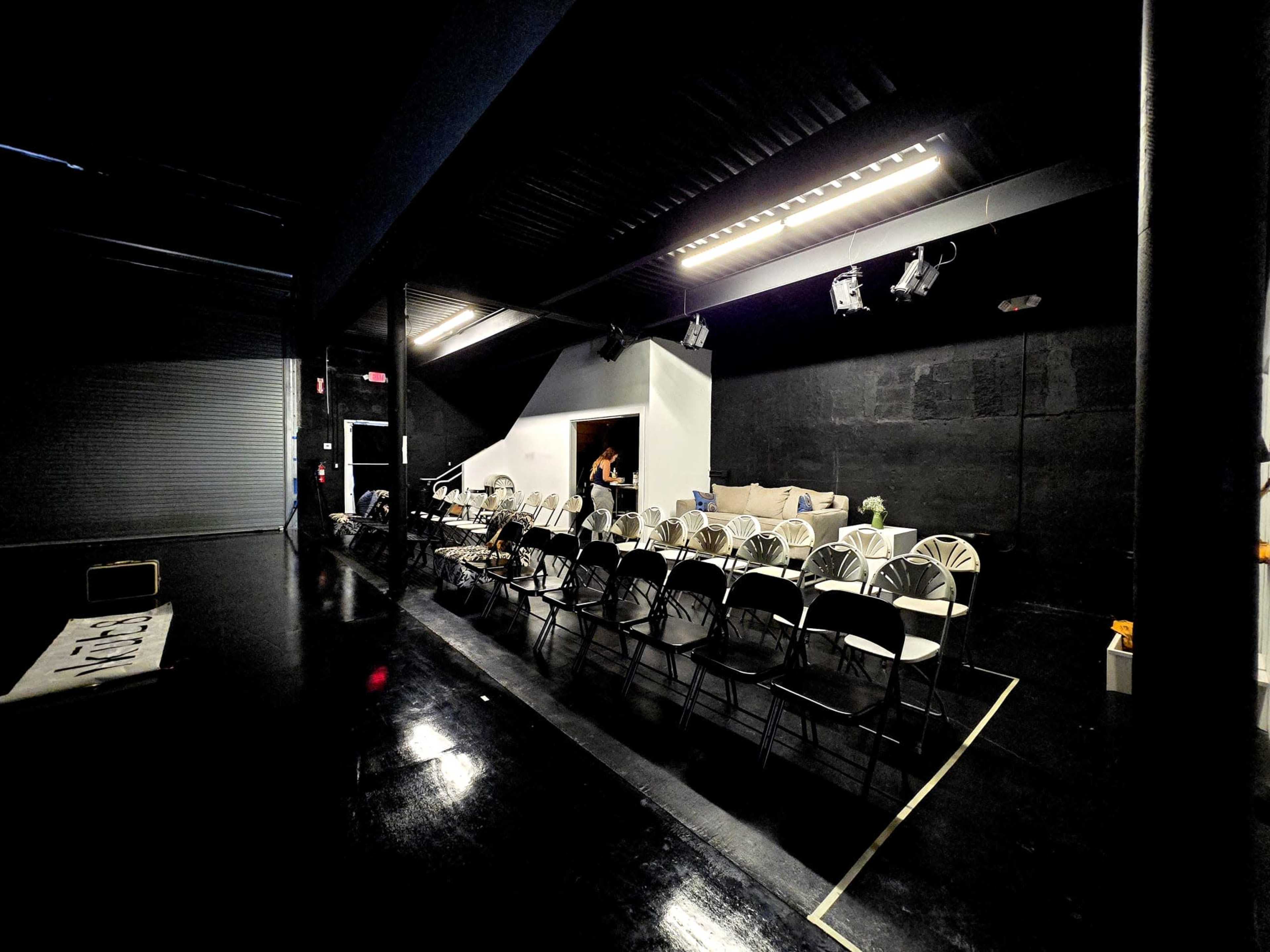 The image shows a dimly lit performance space with rows of black folding chairs arranged in front of a plain white wall featuring a large artwork.