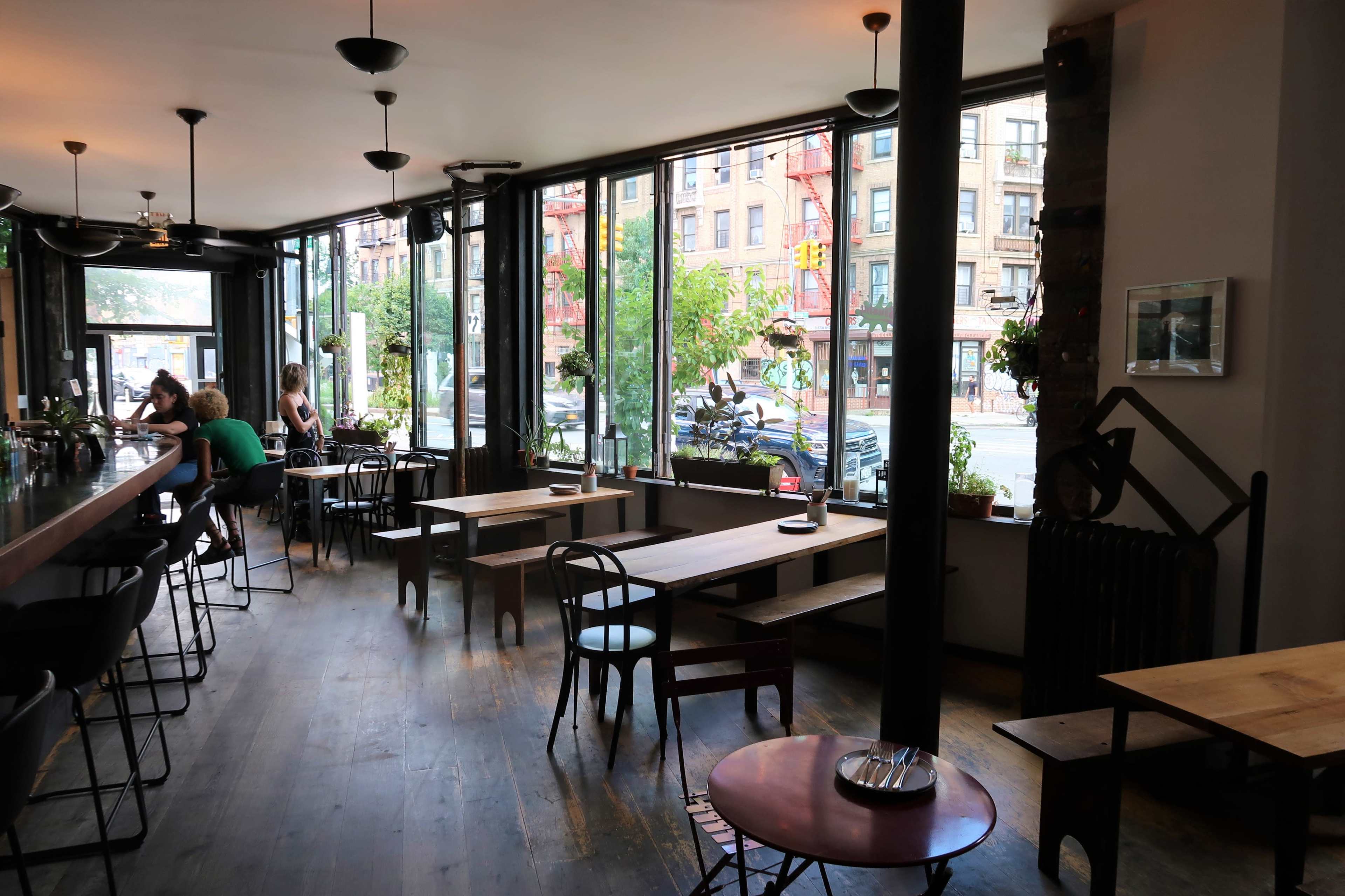 A modern cafe with large windows, wooden tables, and chairs, featuring a view of a city street outside.