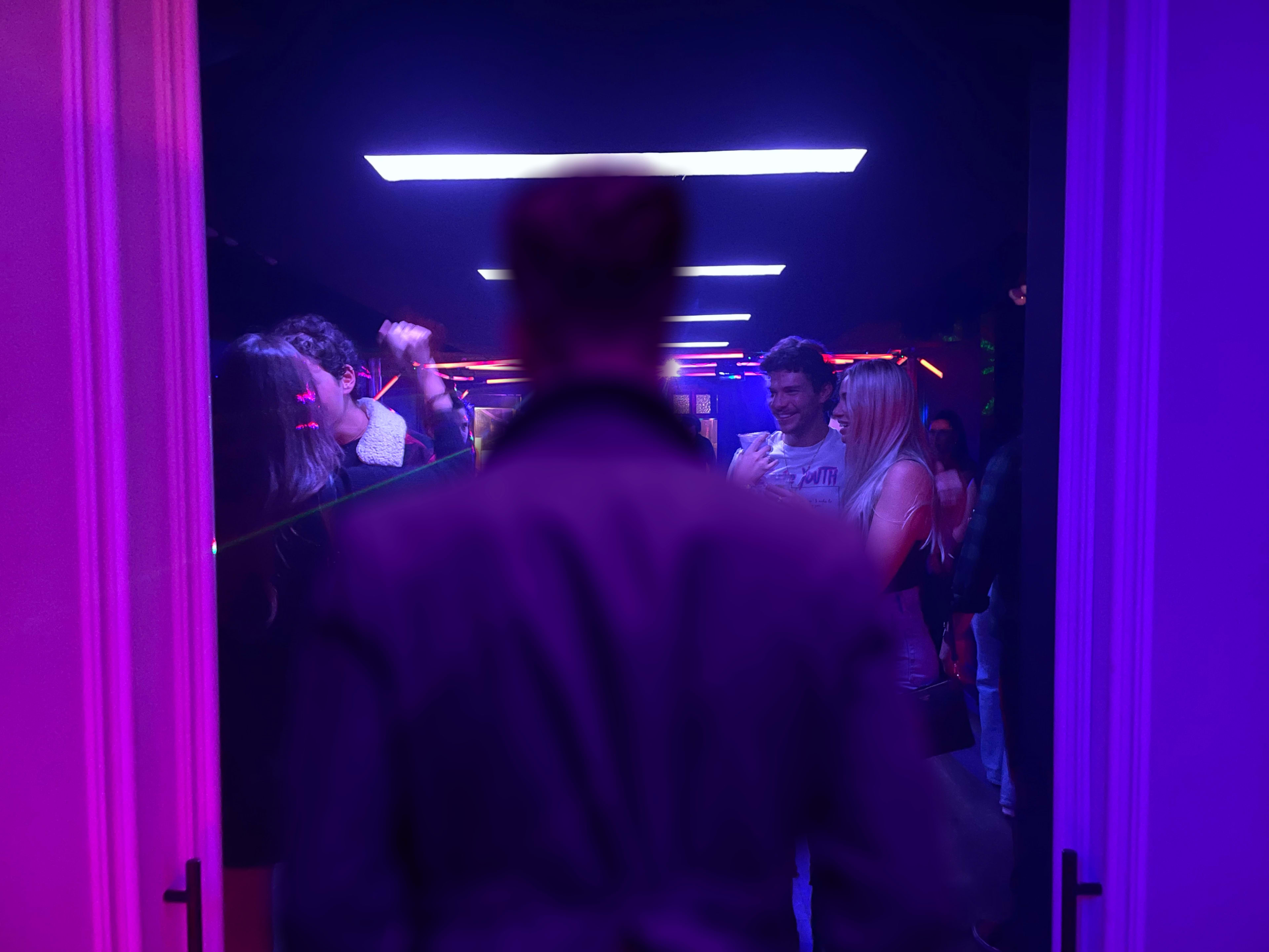 A person stands in a doorway looking into a dimly lit space filled with people dancing under colorful lights.