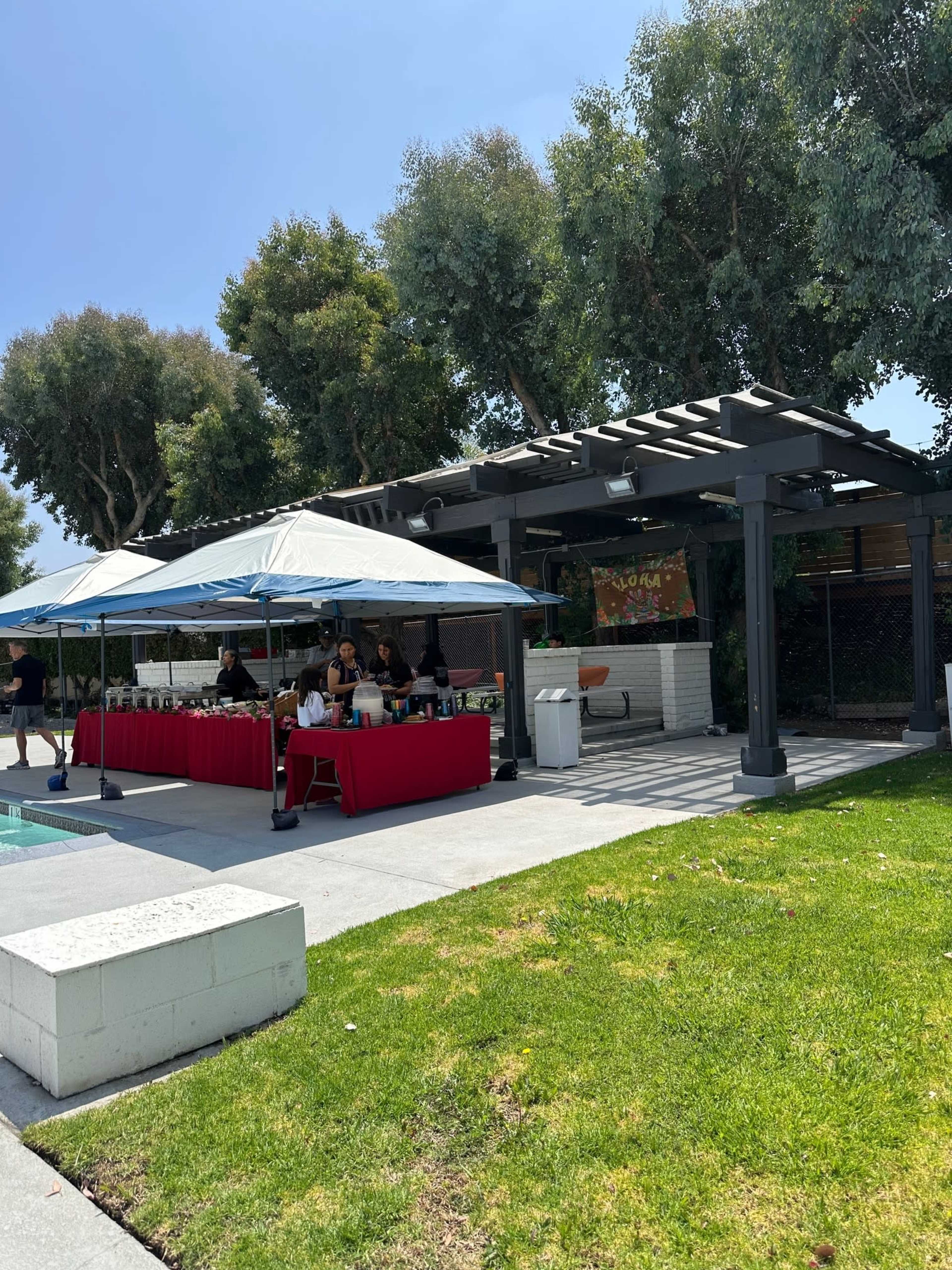 A shaded outdoor area with a red tablecloth set up for an event features a group of people under a blue tent.
