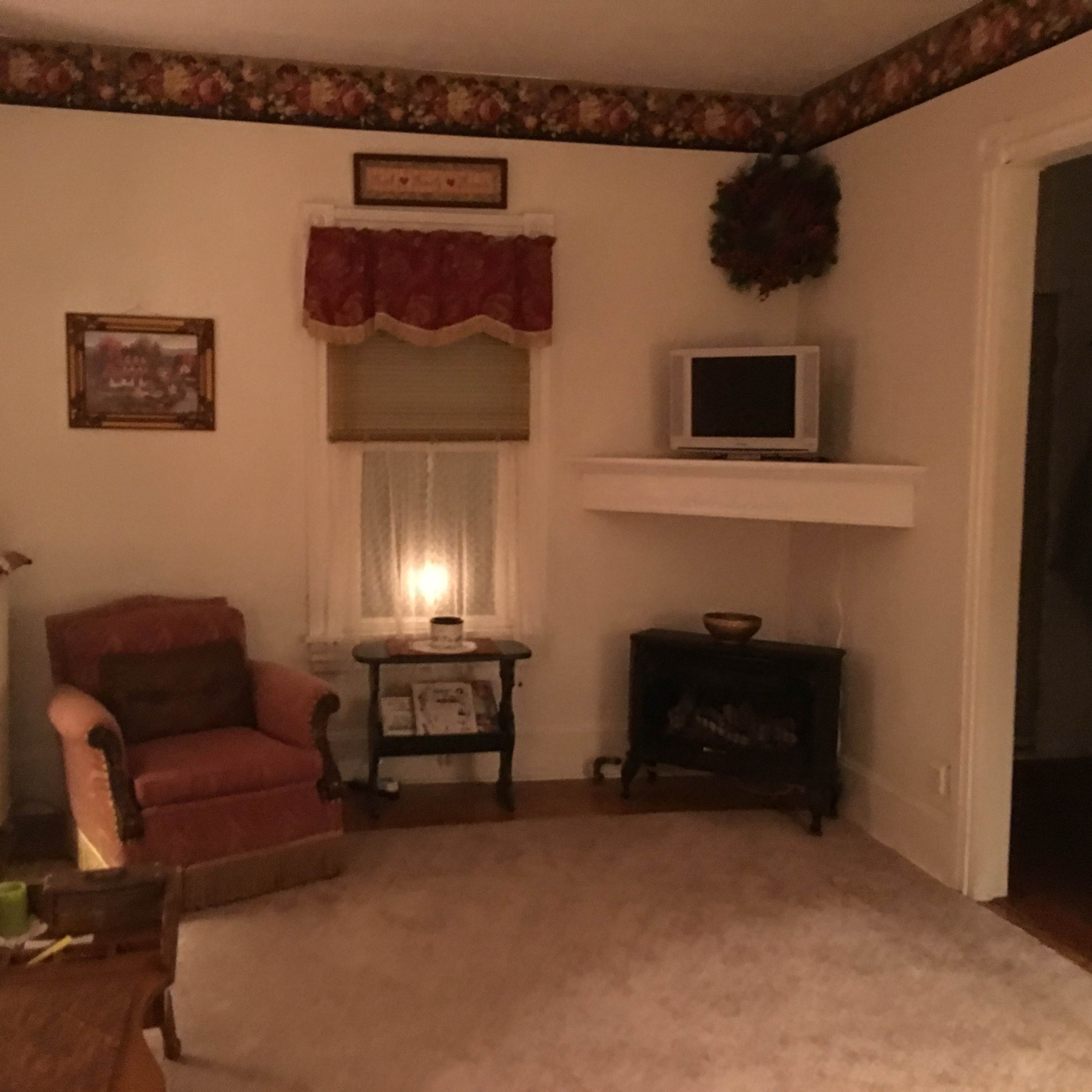 A cozy living room features a pink armchair, a small table with a candle, and a television mounted above an electric fireplace.