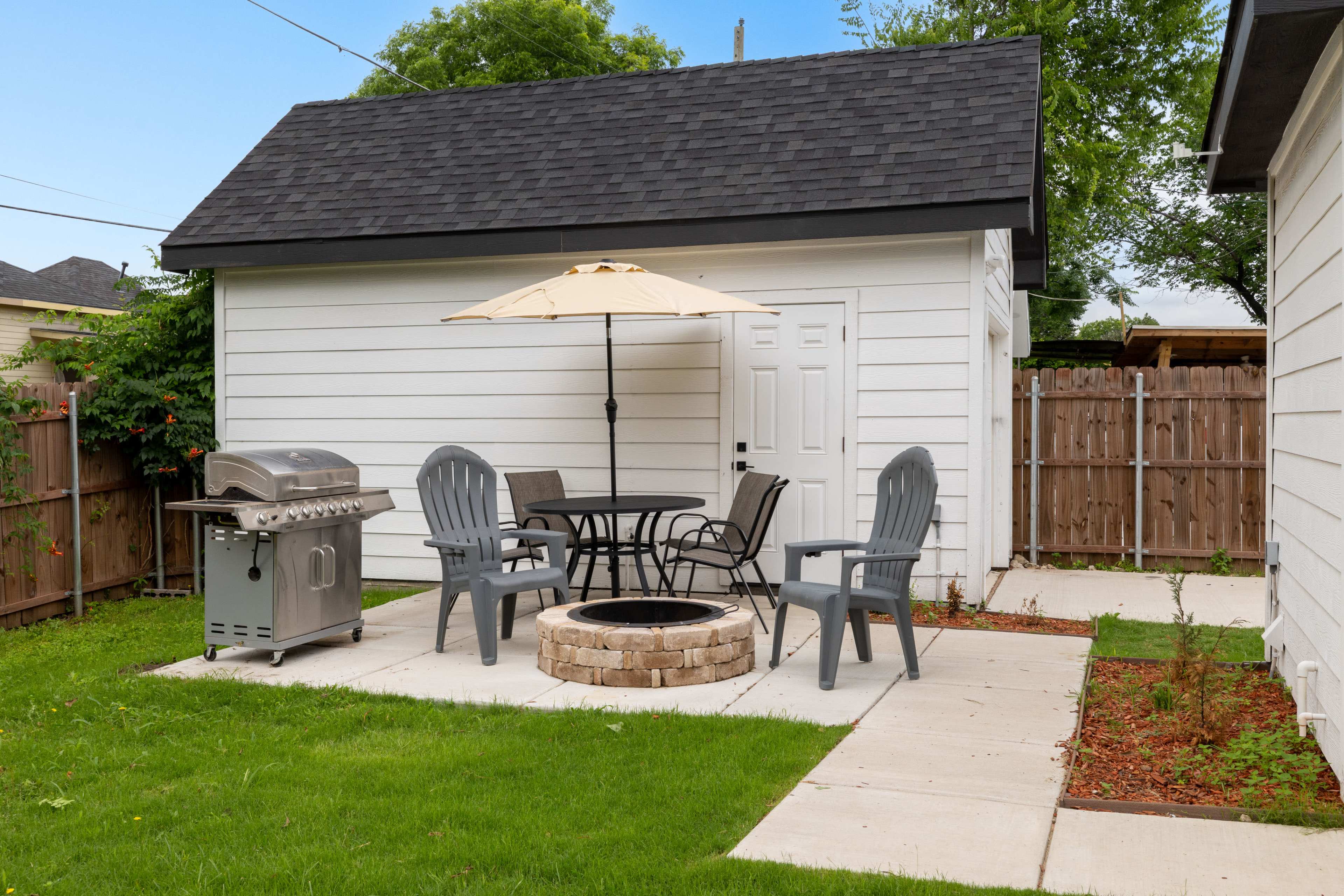 A backyard patio features a table with chairs, an umbrella, a barbecue grill, and a circular stone fire pit.