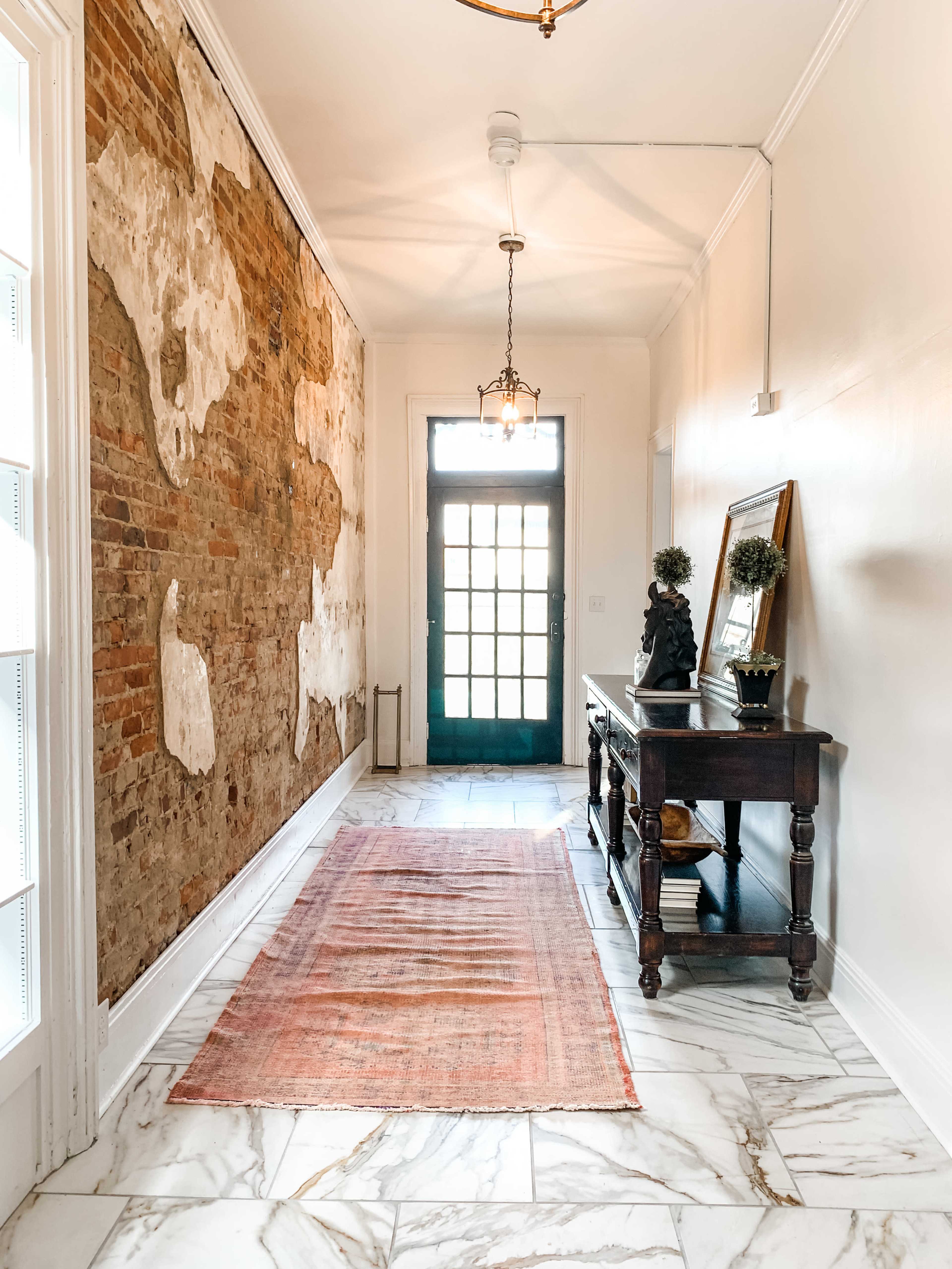 A long hallway featuring exposed brick walls, a wooden console table, a decorative statue, and a patterned rug leading to a door with glass panels.