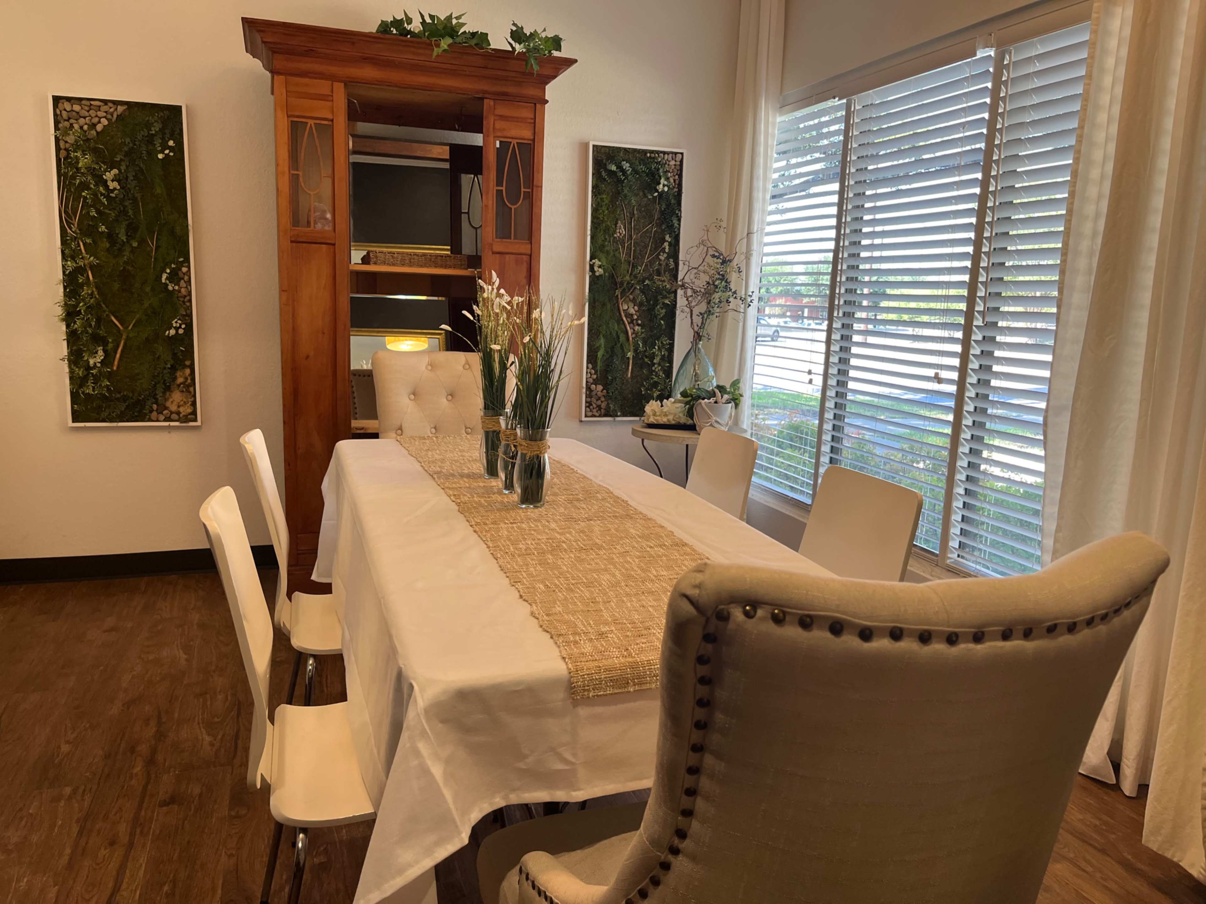 A dining area features a long table set with a table runner, surrounded by white chairs and a tufted armchair, complemented by shelves and framed greenery on the walls.