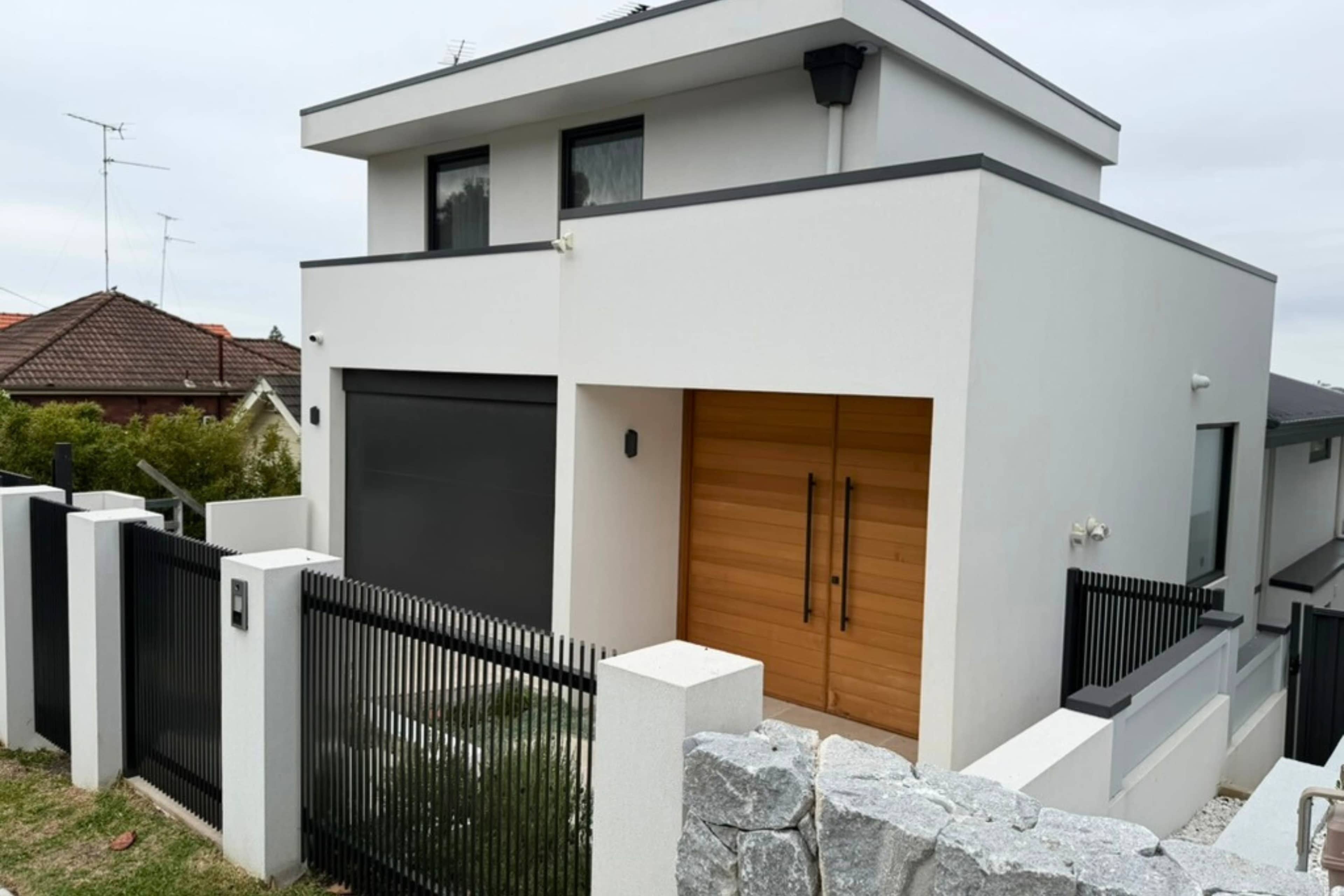 The image shows a modern two-story house with a white exterior, a black garage door, and a wooden front door framed by a black fence.