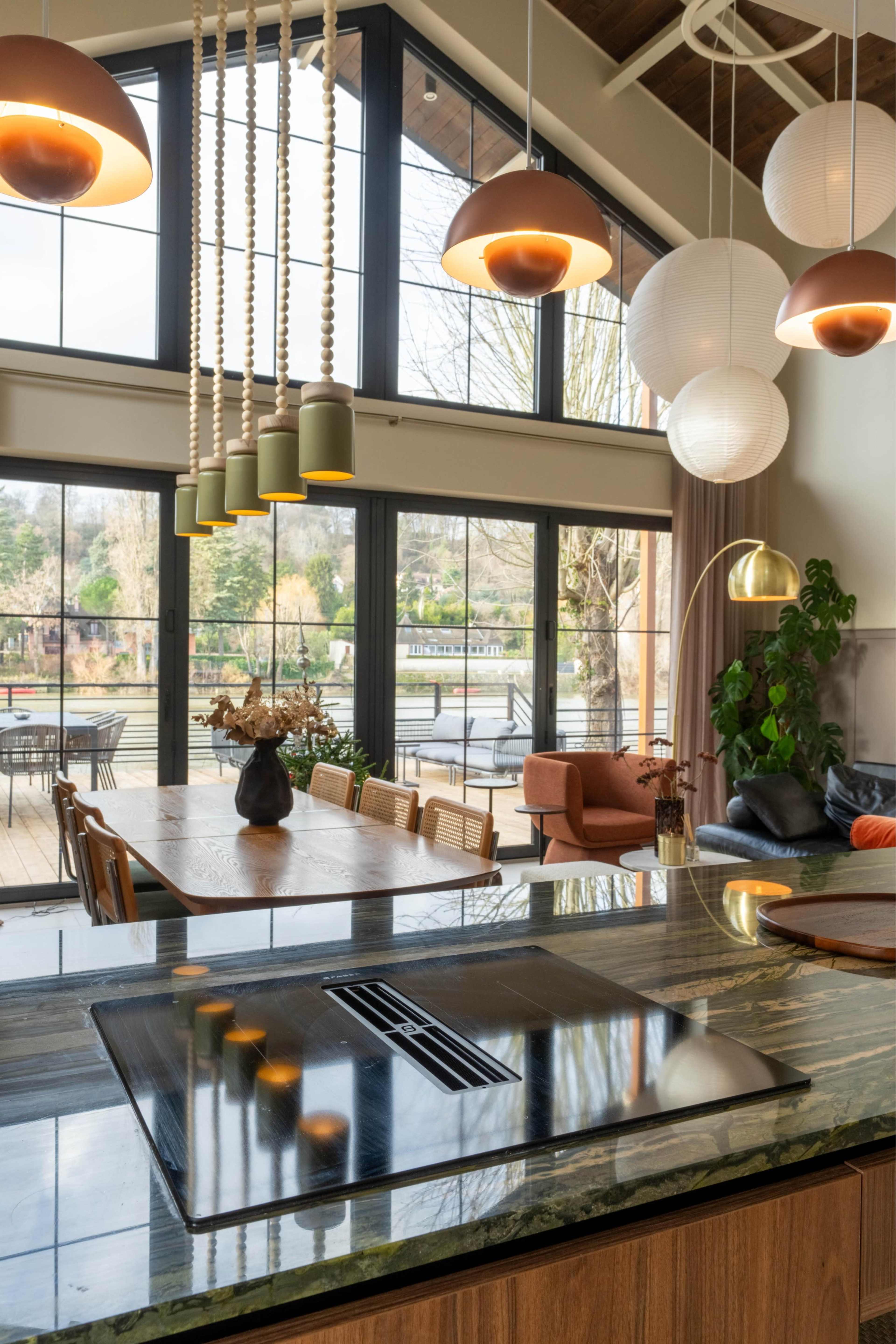 The image shows a spacious kitchen and dining area with modern lighting fixtures and large windows that overlook an outdoor deck and garden.