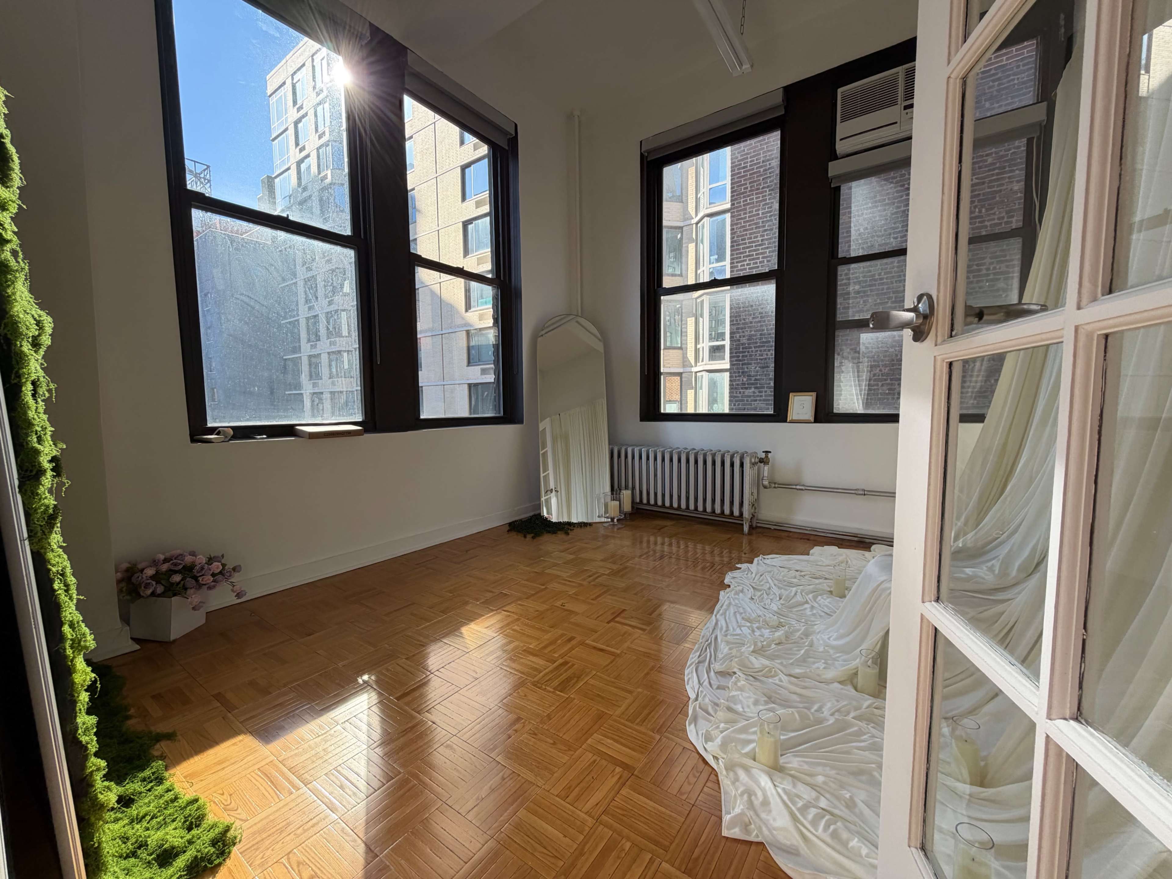 A bright room features large windows, a wooden floor, and white drapery arranged on the ground, with greenery and a mirror in the corner.