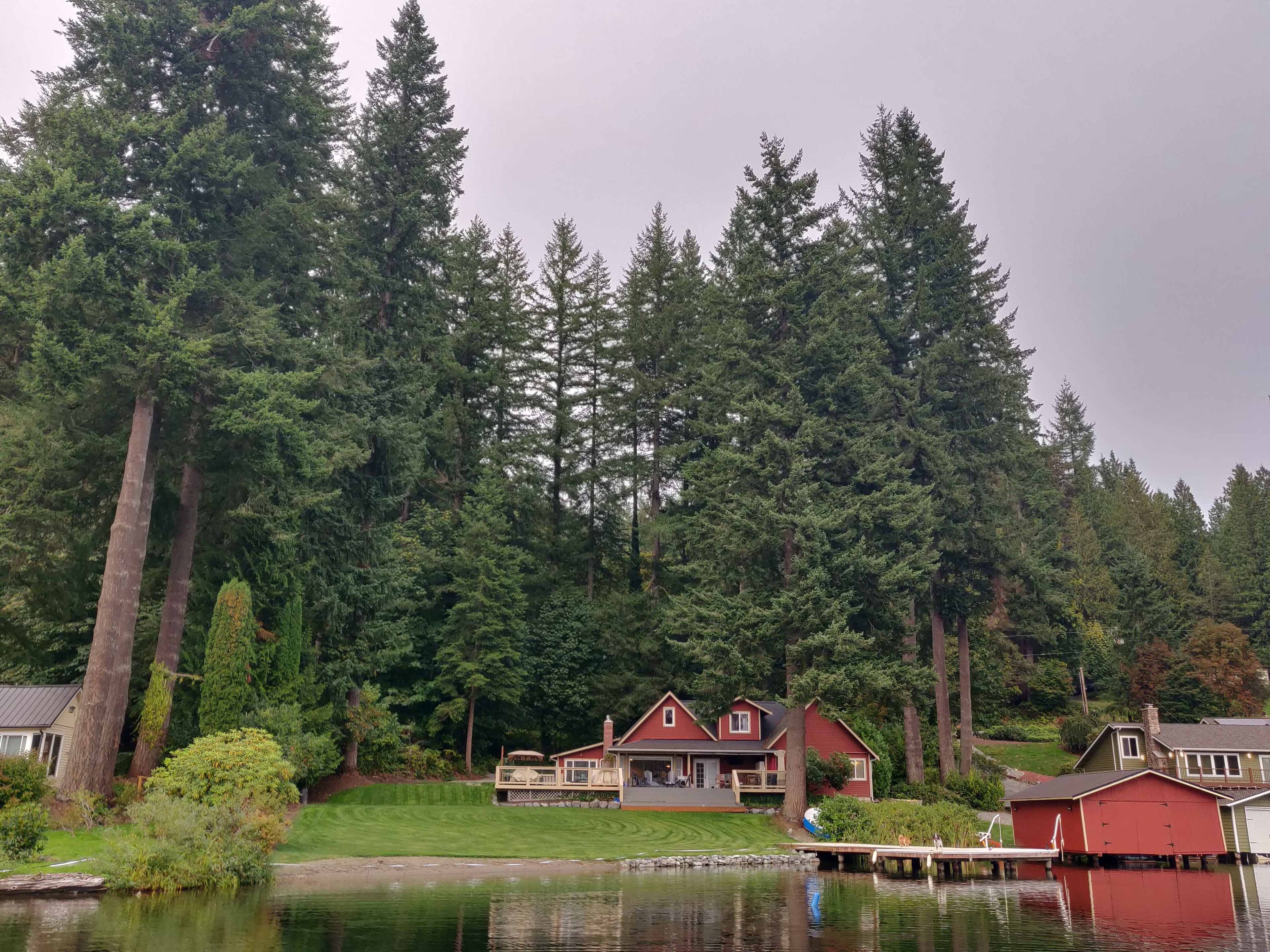 A red house with a deck sits near the water, surrounded by tall evergreen trees and a small boathouse on the shore.