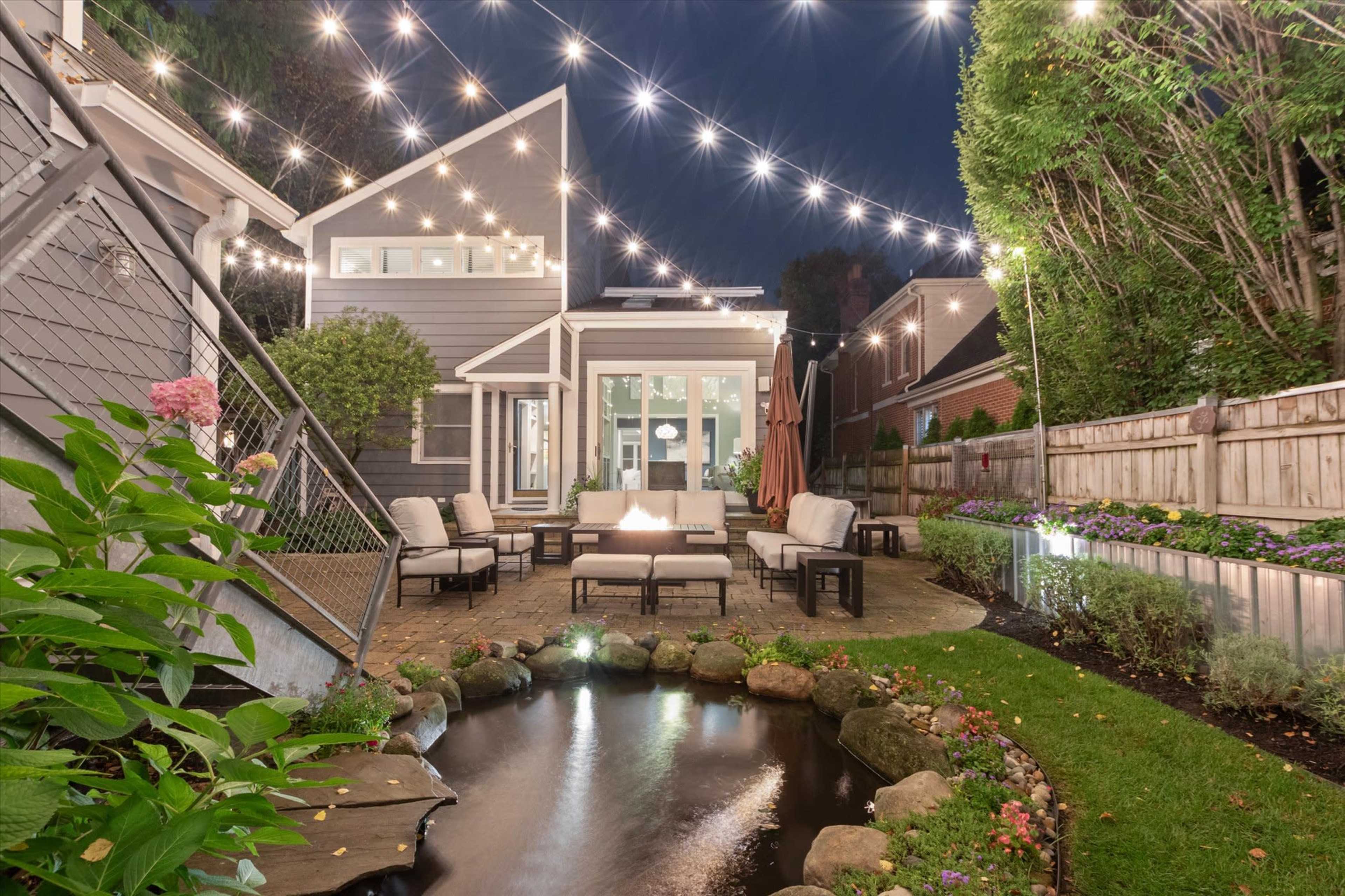 A well-lit backyard features a seating area with a fire pit, surrounded by a pond and neatly arranged landscaping.