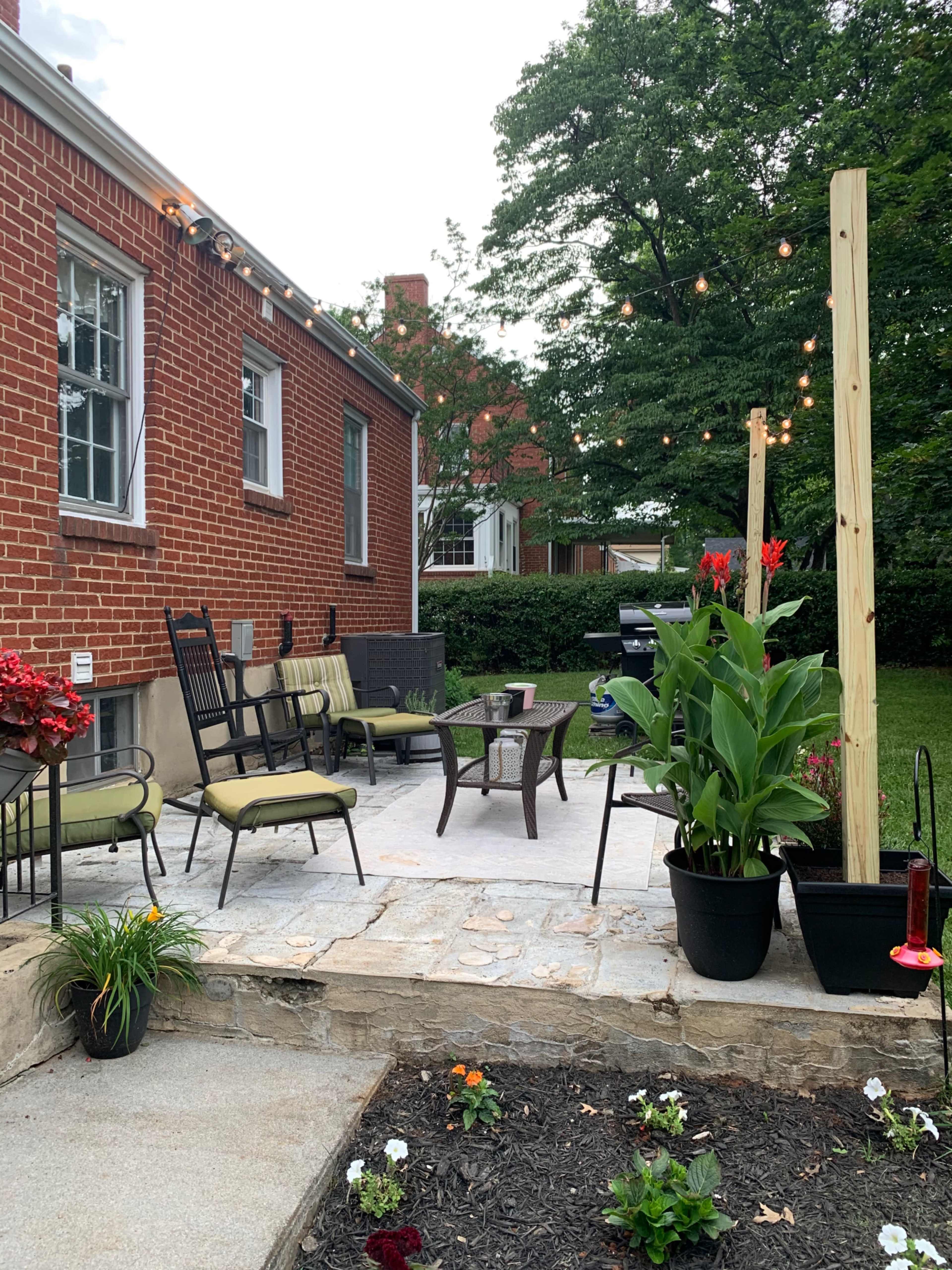 A stone patio area with outdoor seating, potted plants, and string lights attached to the wooden posts, set against a brick wall and greenery.