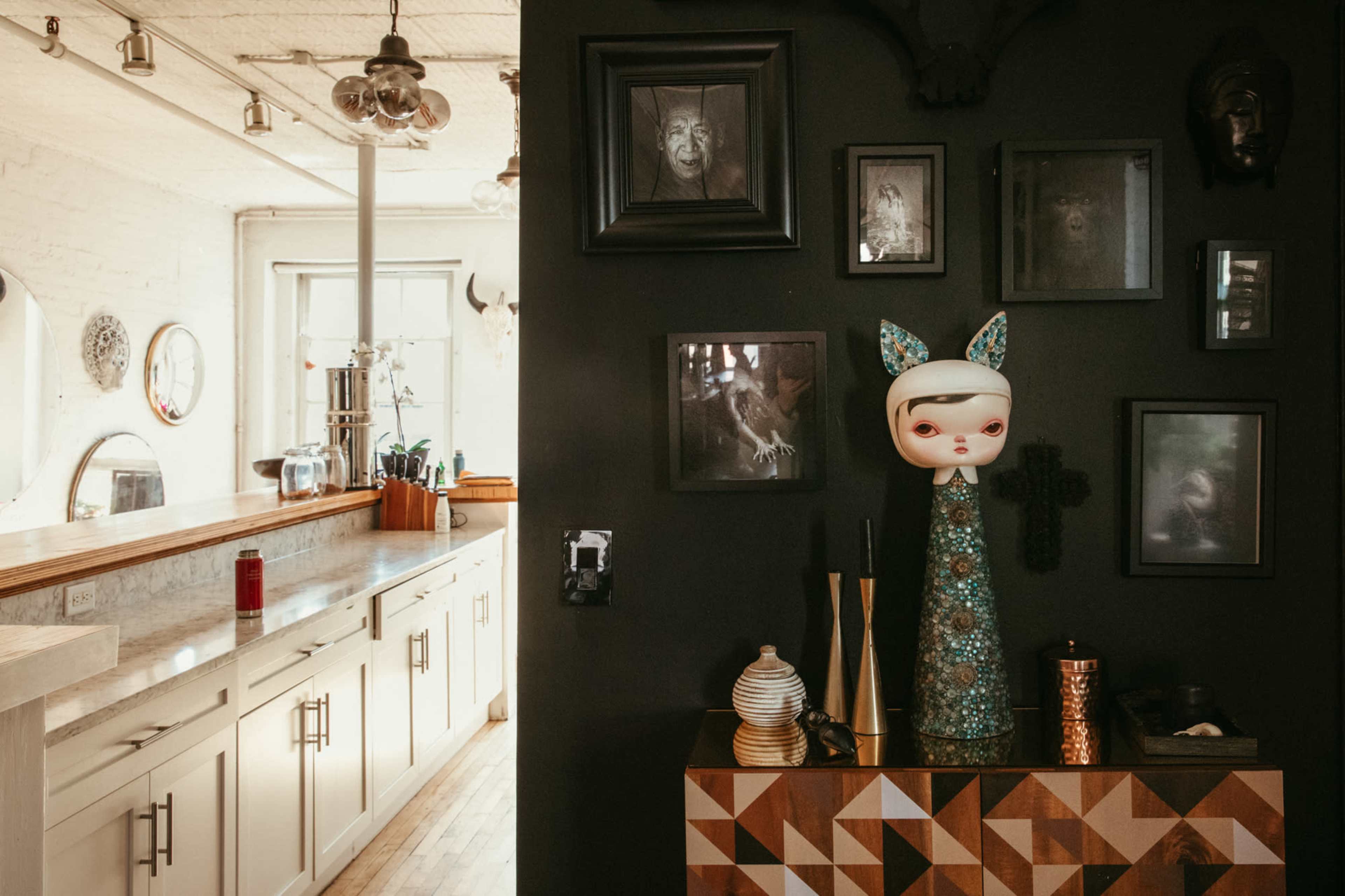 A stylish interior with a dark wall displaying framed art and a decorative figure on a geometric-patterned cabinet, complemented by a bright kitchen area in the background.