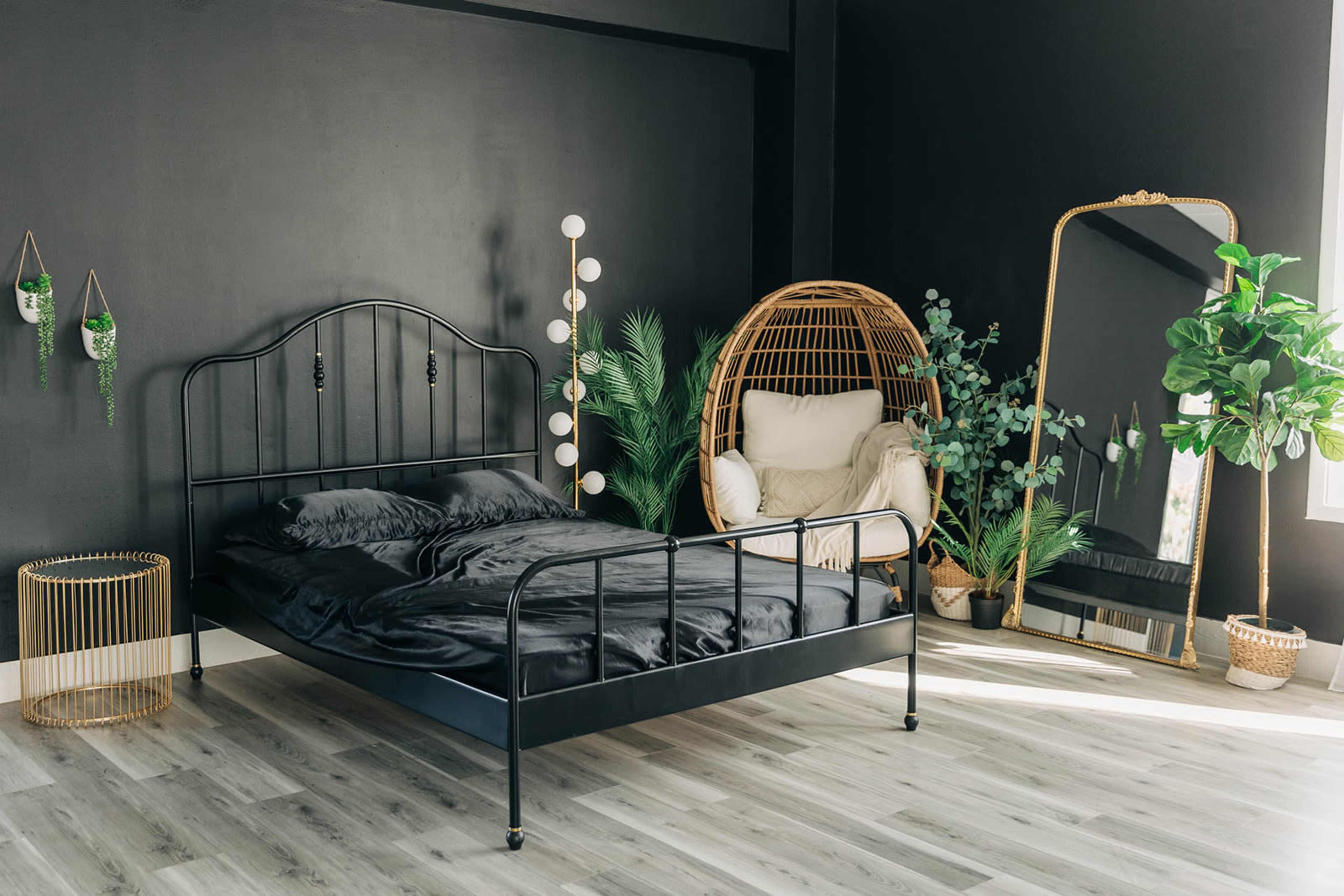 The image shows a minimalist bedroom featuring a black metal bed frame with dark bedding, a large mirror, a rattan chair, and various plants against a black wall.