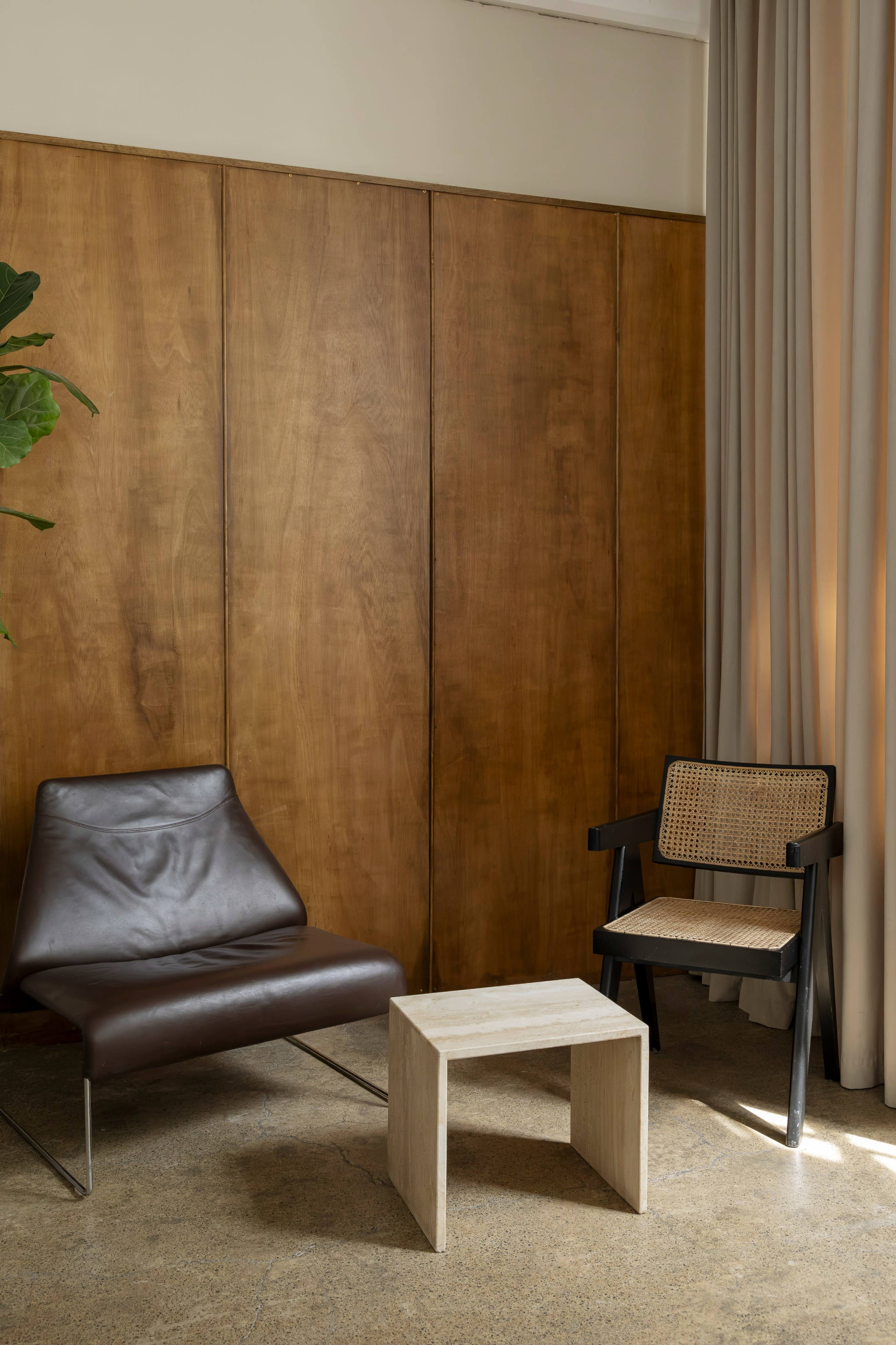 The image shows a simple seating area featuring a modern brown lounge chair, a wooden side table, and a black woven chair against a backdrop of wood-paneled walls and light curtains.