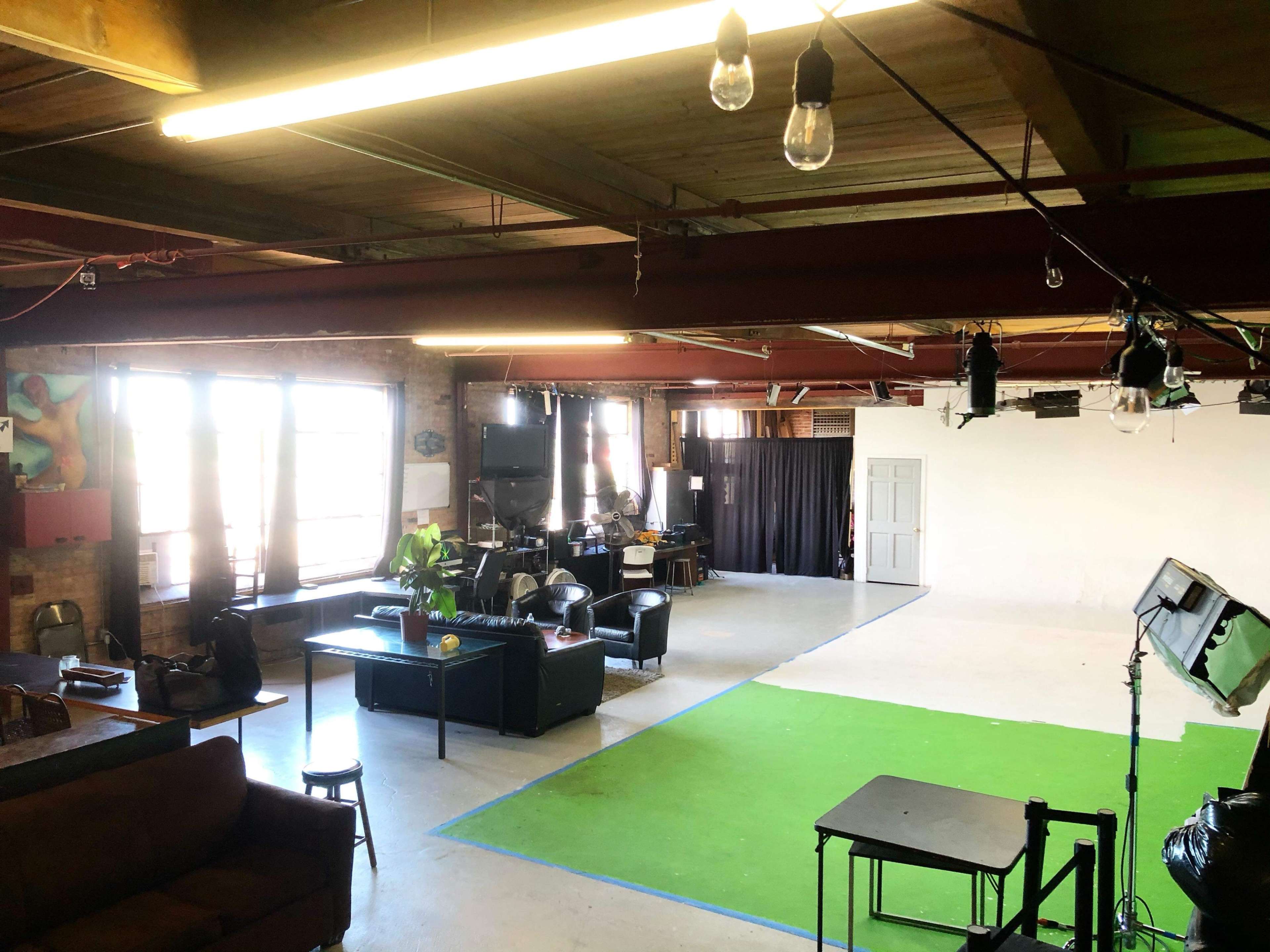 An industrial-style studio space with large windows, a green carpet area, and various furniture and equipment arranged throughout.