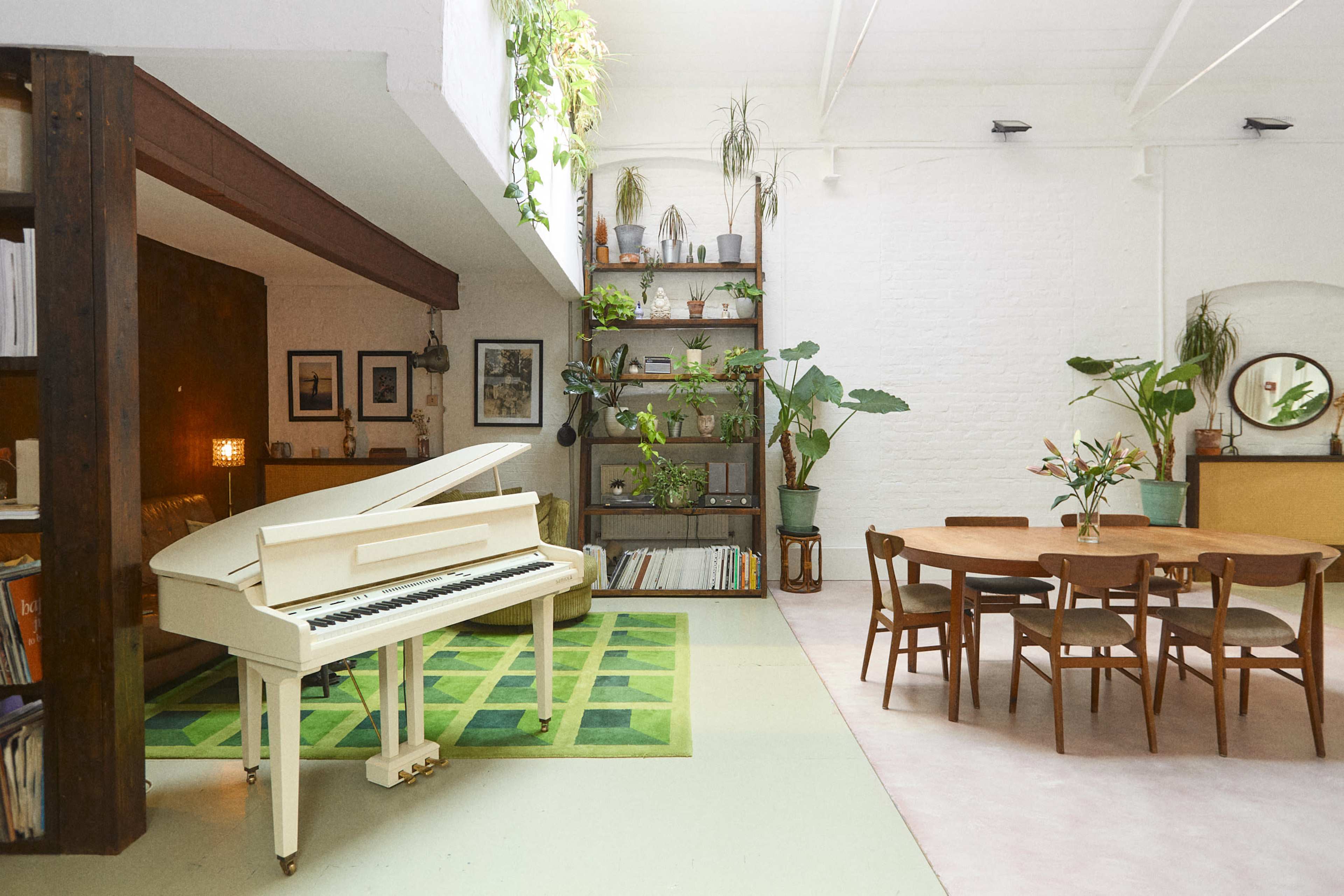 The image features a bright living space with a white piano, a round wooden dining table, and various indoor plants arranged throughout.