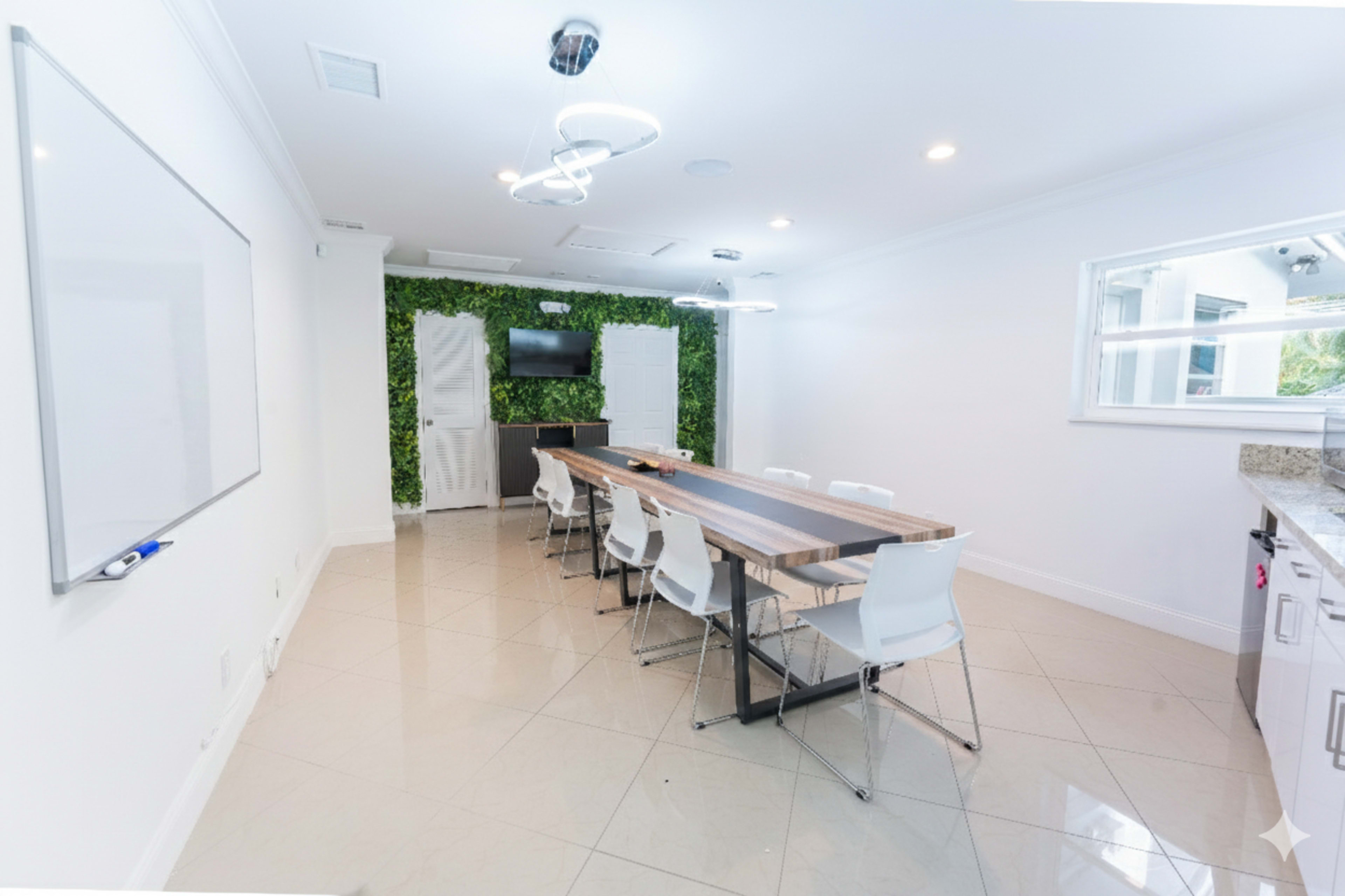 The image depicts a modern conference room with a long wooden table surrounded by white chairs, a green plant wall, and a wall-mounted television.