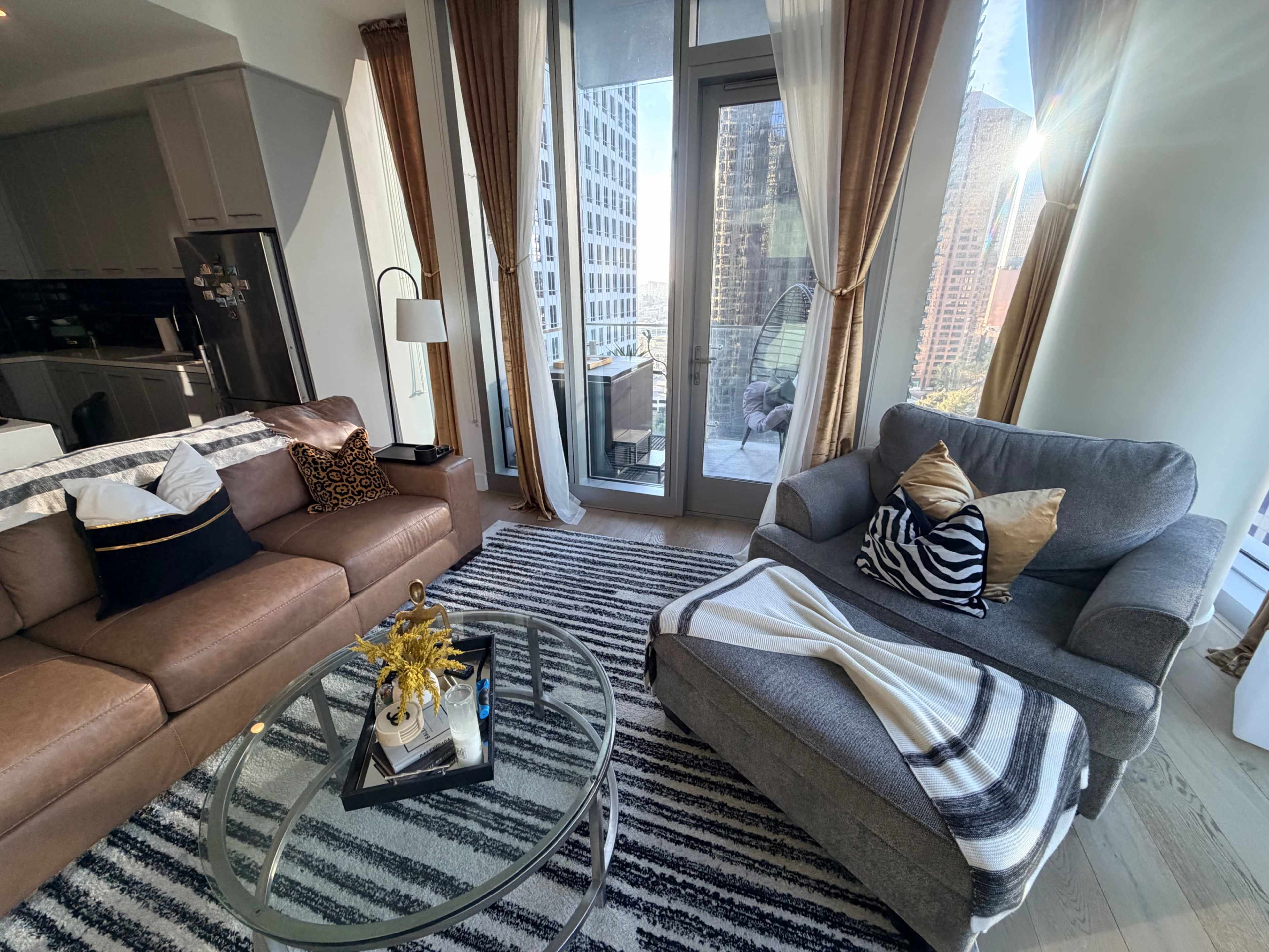 A modern living room features a brown leather sofa and a gray armchair, with large windows showing a cityscape and a patterned rug on the wooden floor.