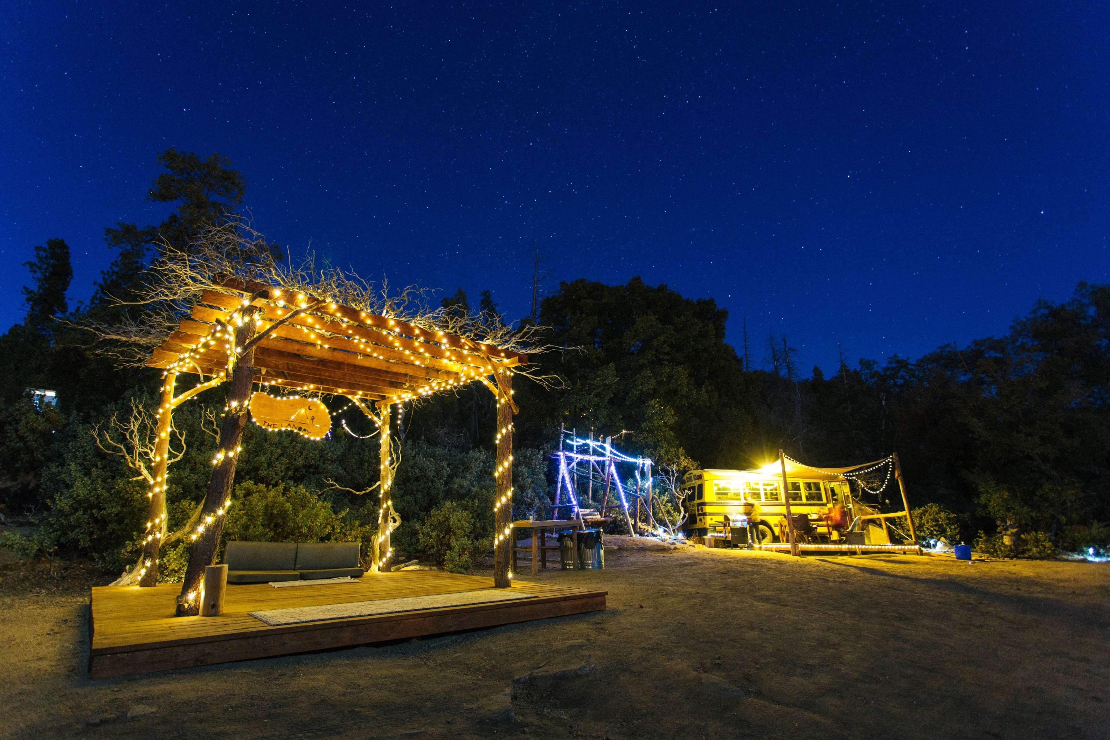 A wooden deck with string lights surrounds a sofa, while a yellow bus is illuminated nearby under a starry night sky.
