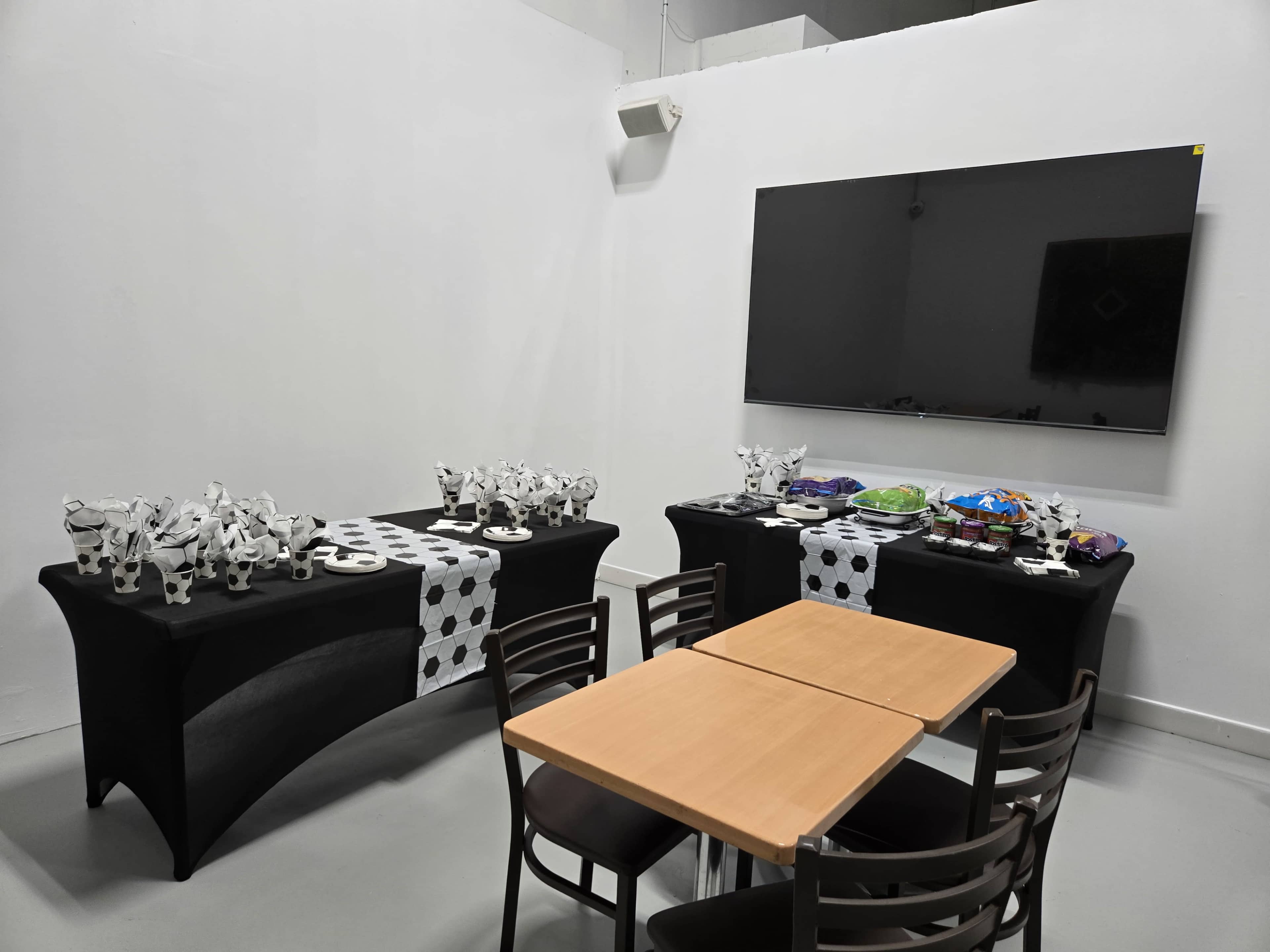 Two tables covered with black cloths and decorated with black-and-white patterns are set up in a room, featuring snacks and drinks, while a large television screen is mounted on the wall.