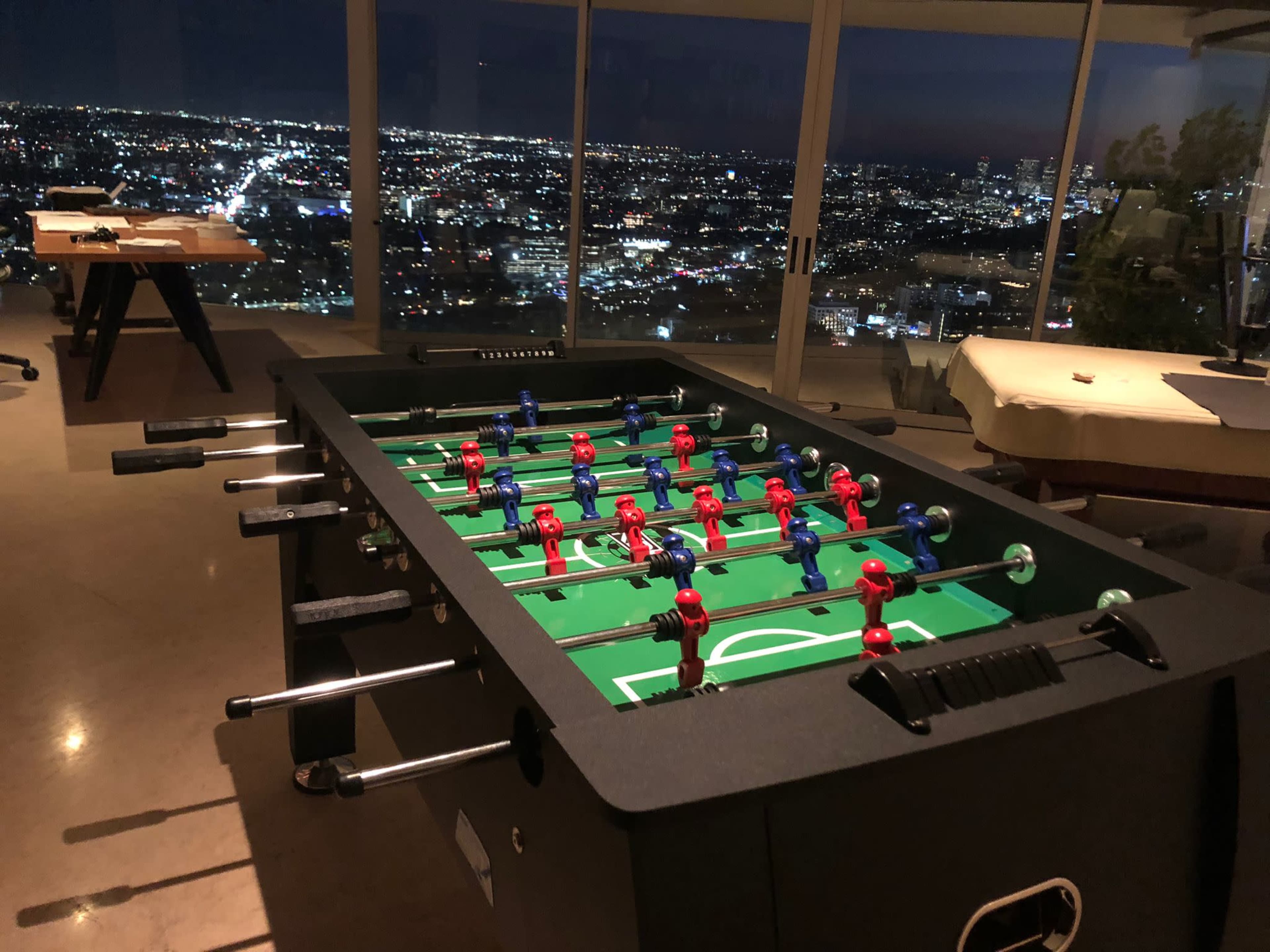 A foosball table sits in a sleek room with large windows overlooking a city skyline at night.