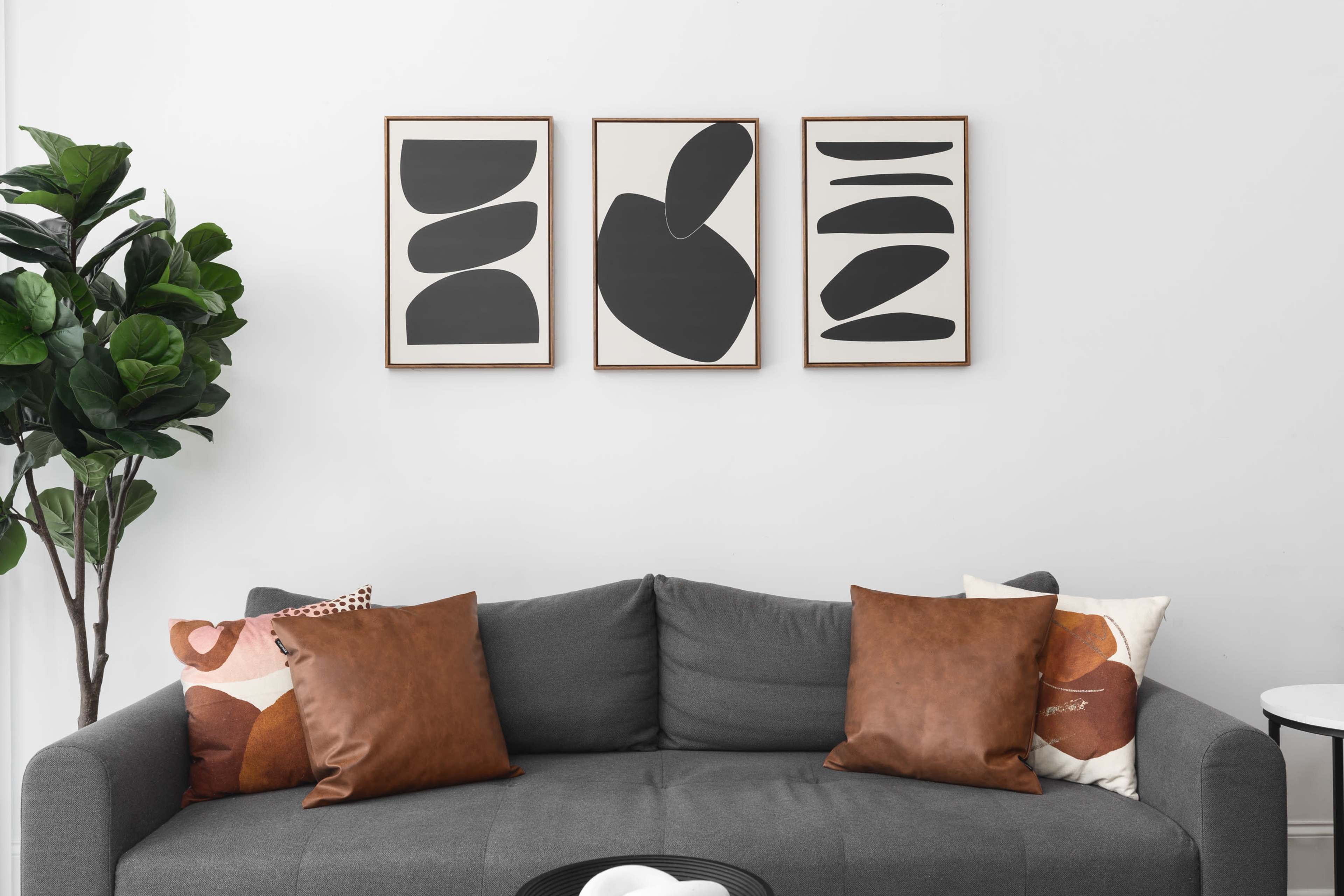 A modern gray couch with brown and patterned pillows is positioned against a white wall adorned with three abstract black and white framed artworks.